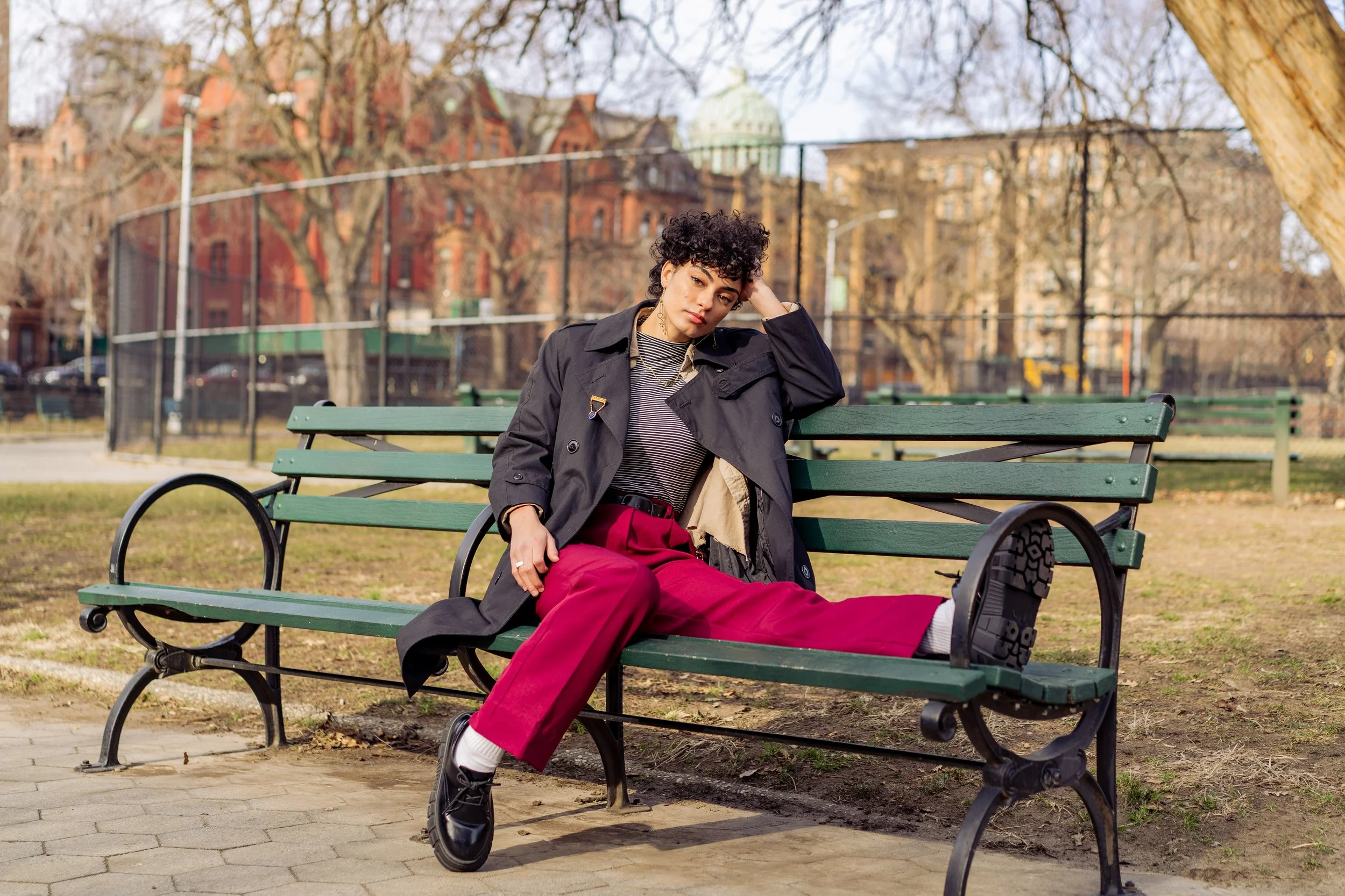 A woman with short curly black hair sitting on a green park bench in a city park. She is wearing a black trench coat, a striped shirt, bright red pants, gray socks, and black shoes. She appears contemplative, resting her head on her hand, with a back