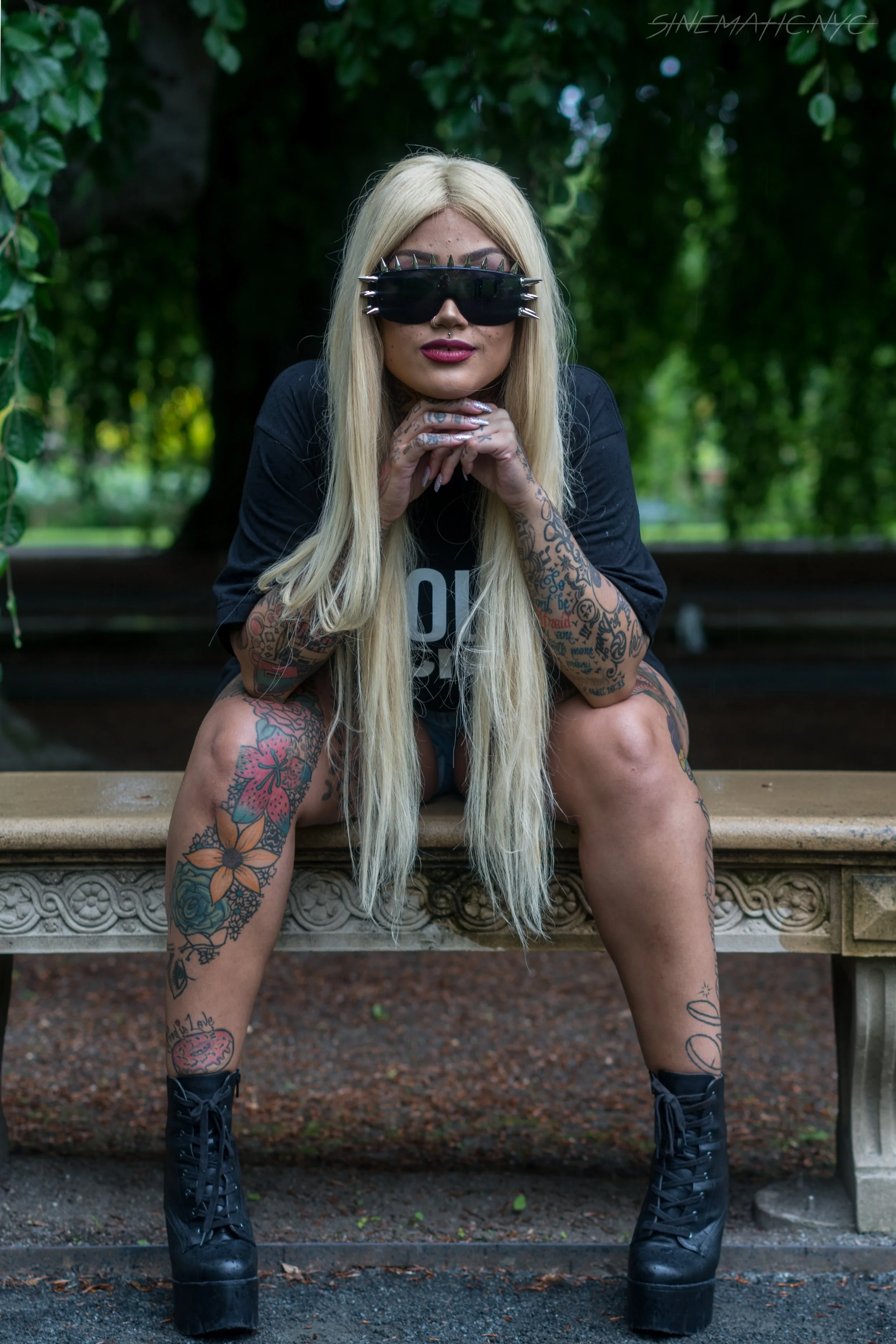 A woman with long platinum blonde hair sitting on a stone bench in a park, wearing large black sunglasses with spikes, a black t-shirt, and black platform boots, with extensive tattoos on her arms and legs.