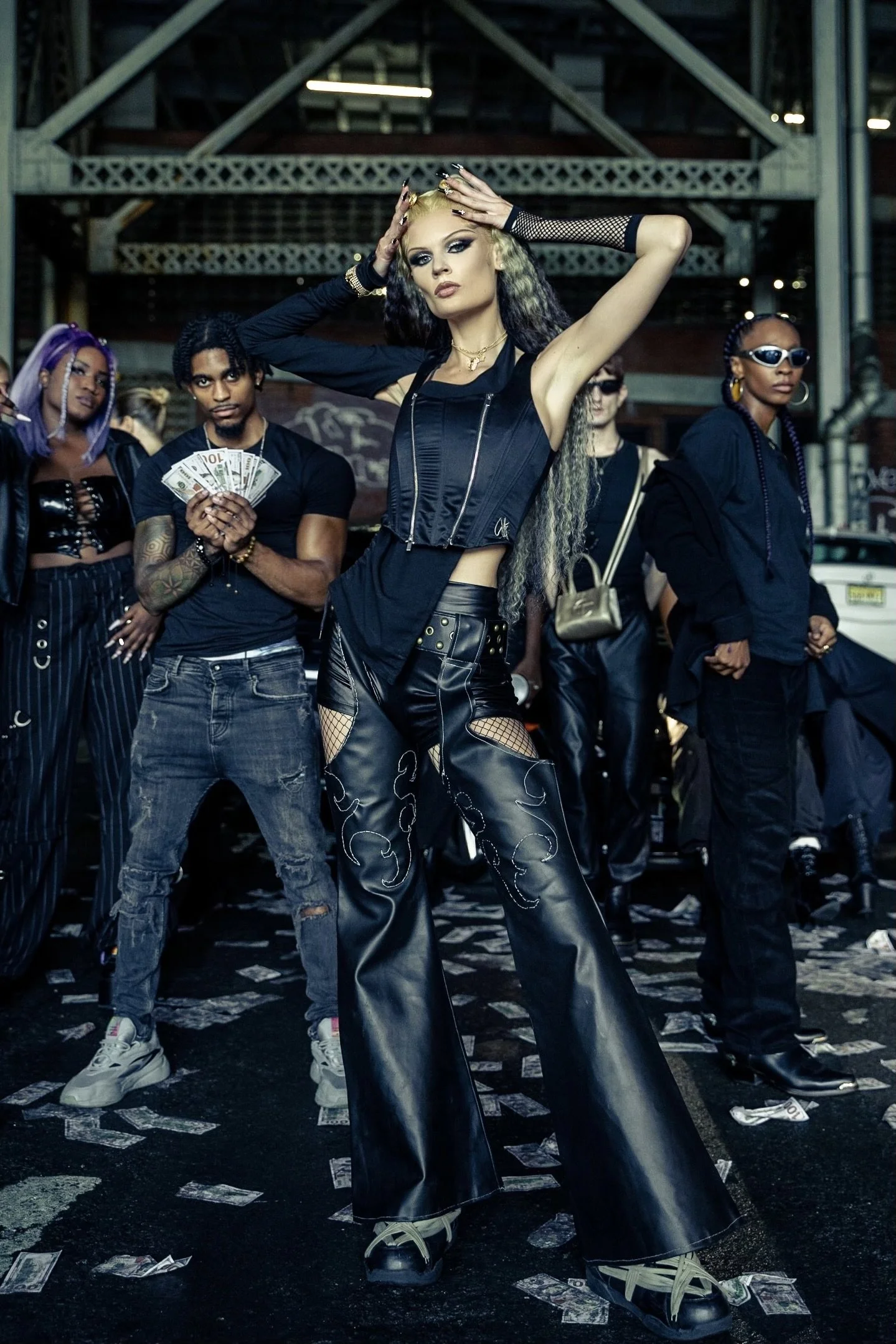 Group of diverse young adults dressed in urban streetwear posing confidently in an industrial setting with dollar bills scattered on the ground.