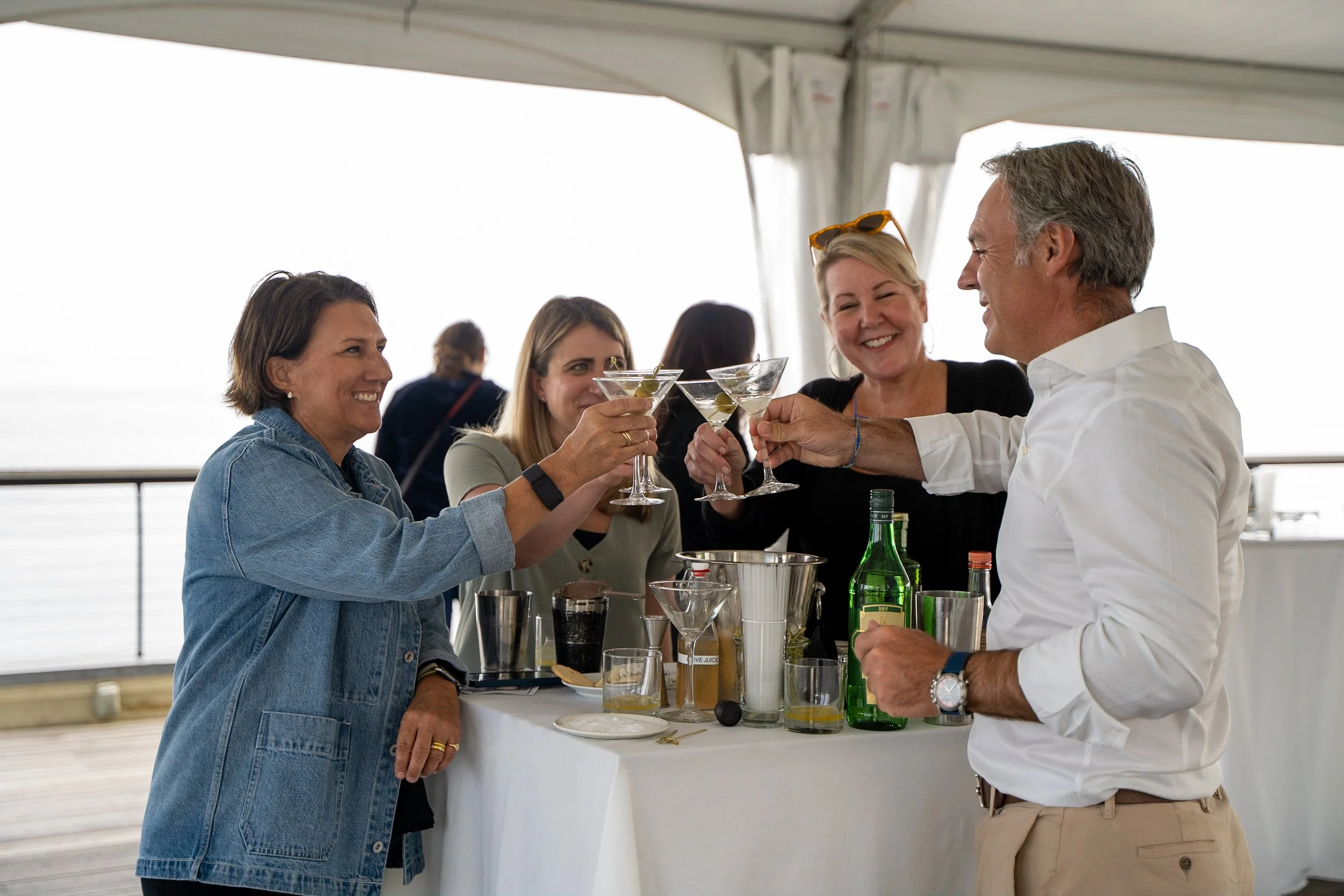 Four adults smiling and toasting with martinis at an outdoor gathering under a tent, with a table of drinks and barware between them.