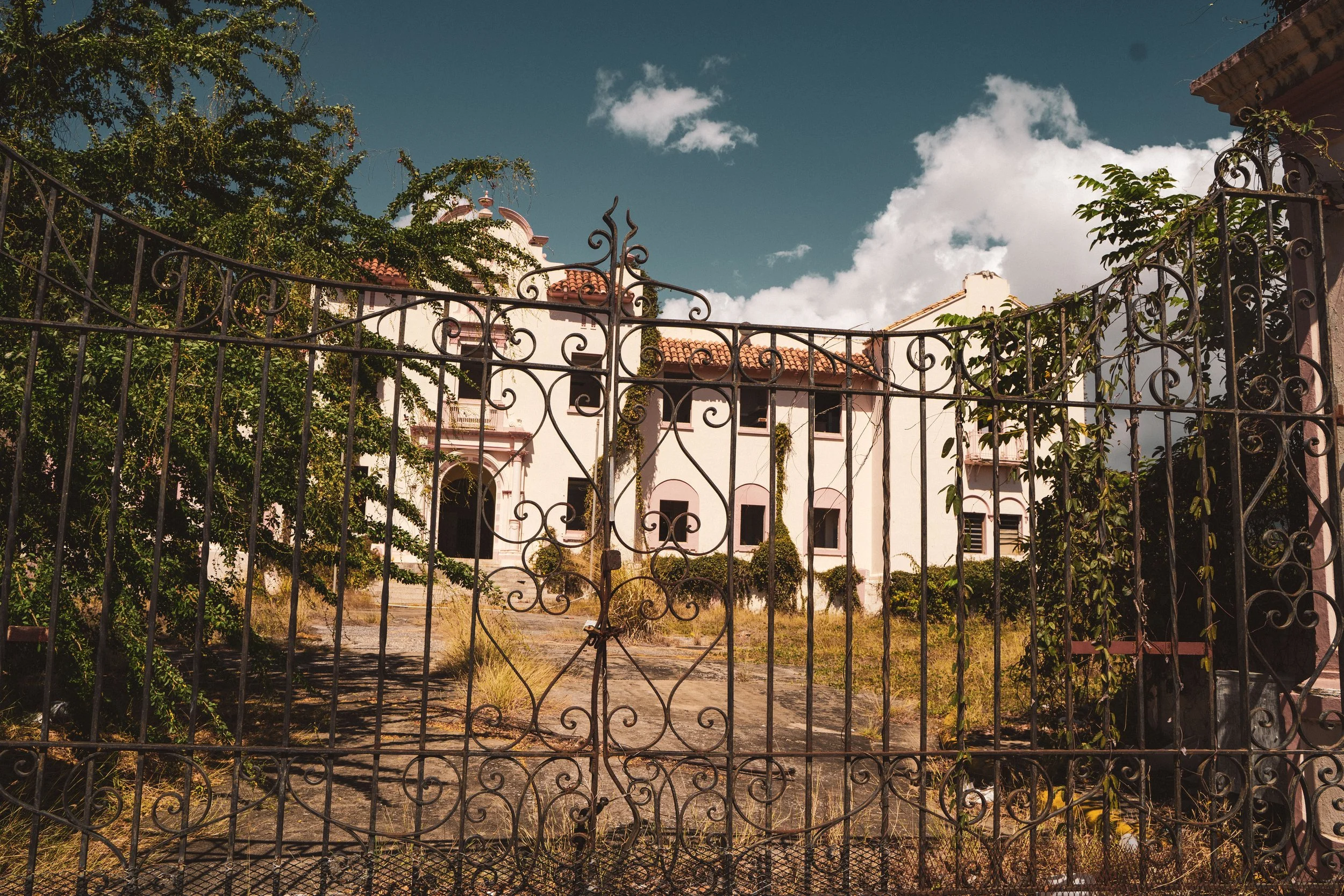 An ornate iron gate in front of an abandoned two-story white building with a red tiled roof, overgrown vegetation, and clear blue sky with a few clouds.