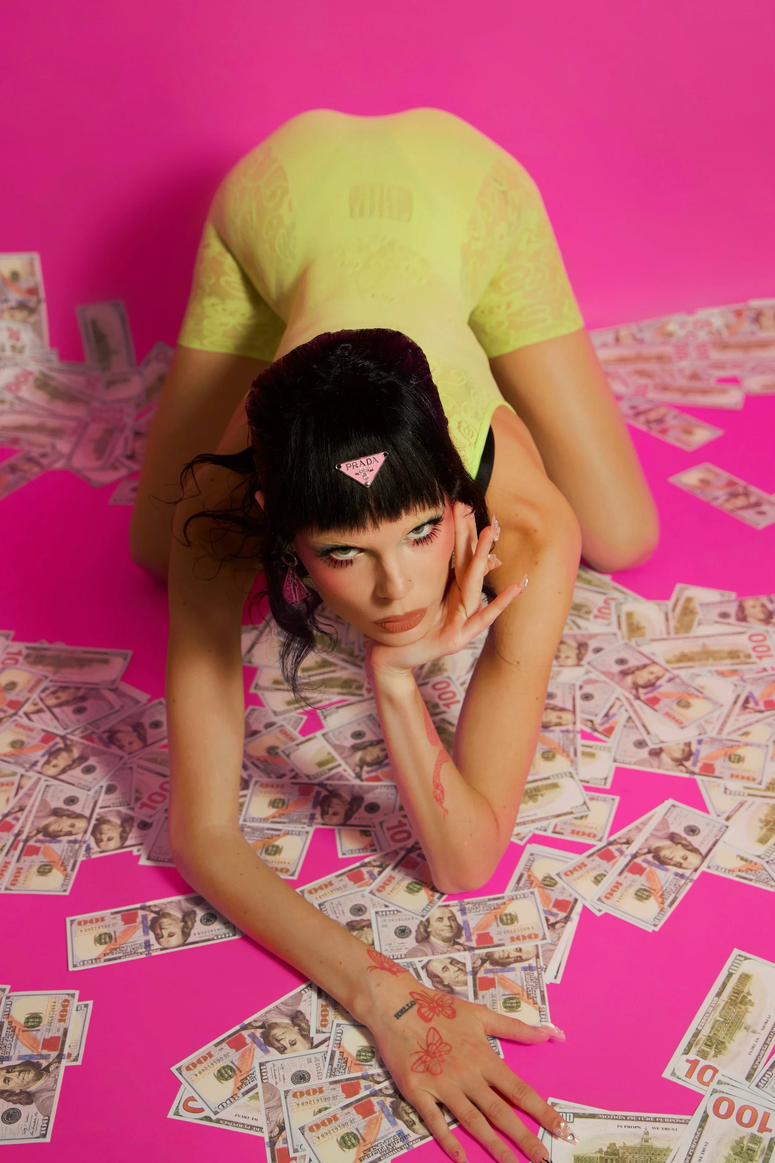 A woman with black hair and tattoos on her arm lying on a pink surface surrounded by scattered hundred-dollar bills, wearing a yellow outfit and a Prada hair accessory.
