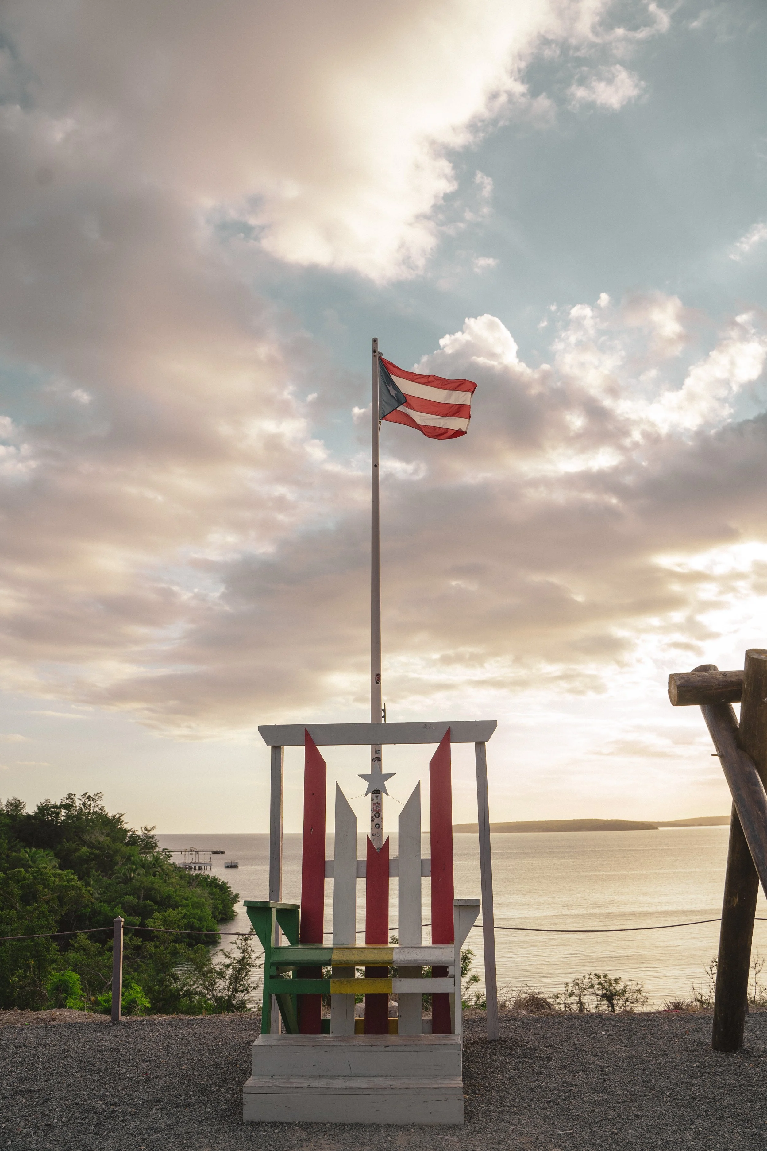 A wooden chair painted in red, white, green, and yellow stripes, placed outdoors near a body of water. An American flag is flying on a pole above the chair, with a background of a cloudy sky during sunset.