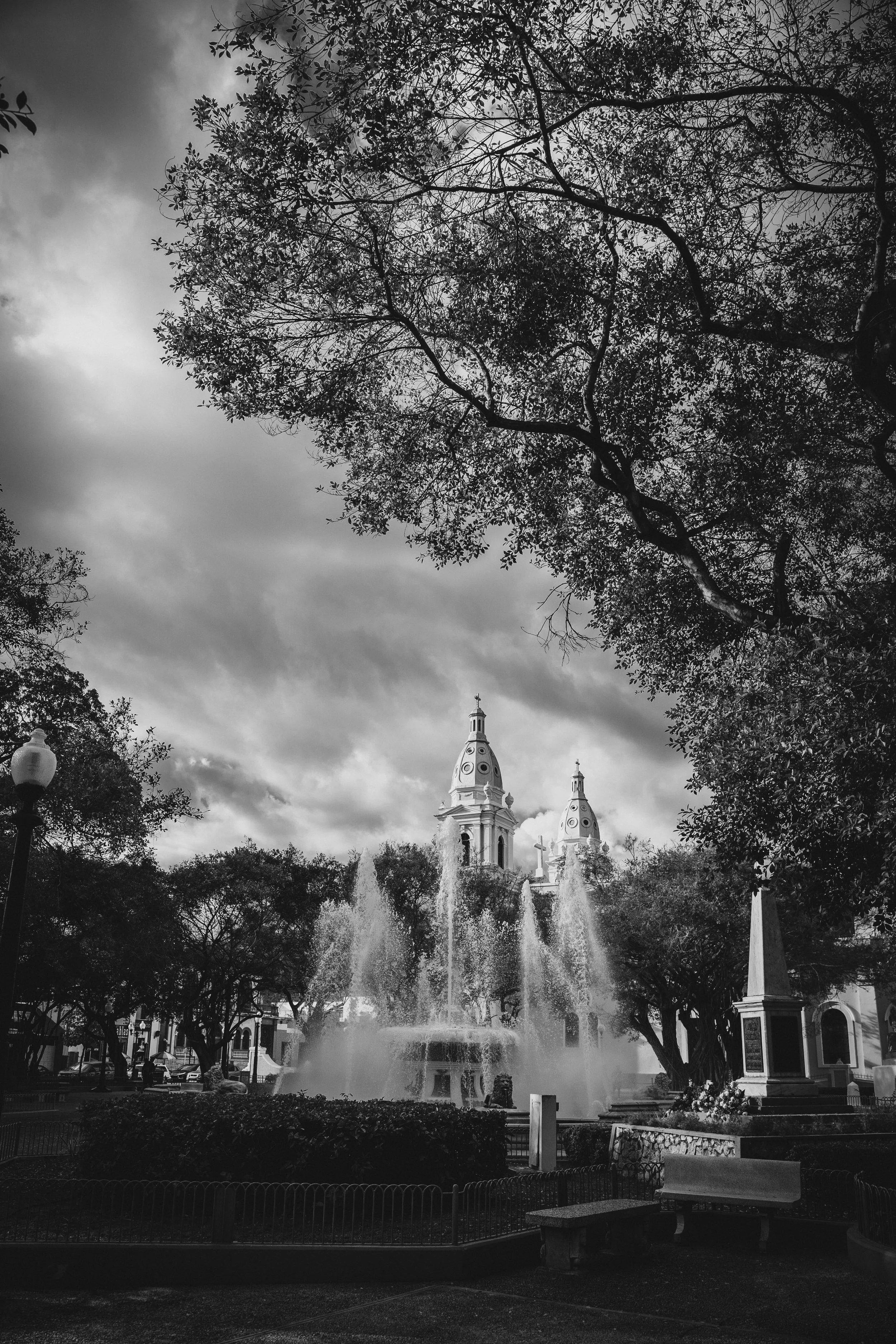 Black and white photo of a park with a fountain, trees, benches, and a church with twin steeples in the background, under a cloudy sky.