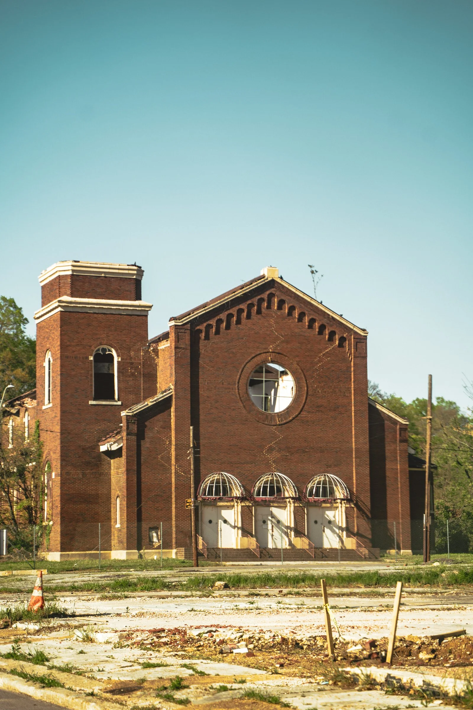 An old, abandoned brick building resembling a church with broken windows and boarded-up doors, surrounded by construction debris and fencing, under a clear blue sky.