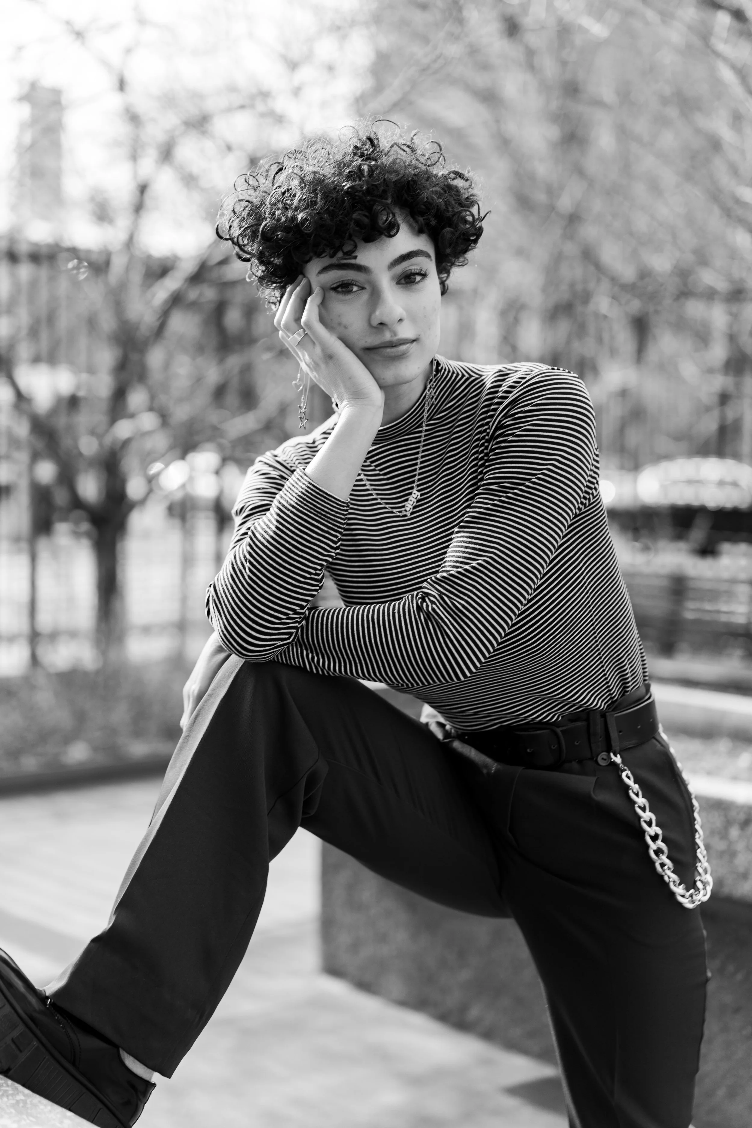A woman with curly hair wearing a striped long sleeve shirt and dark pants, sitting outdoors with trees in the background near Harlem