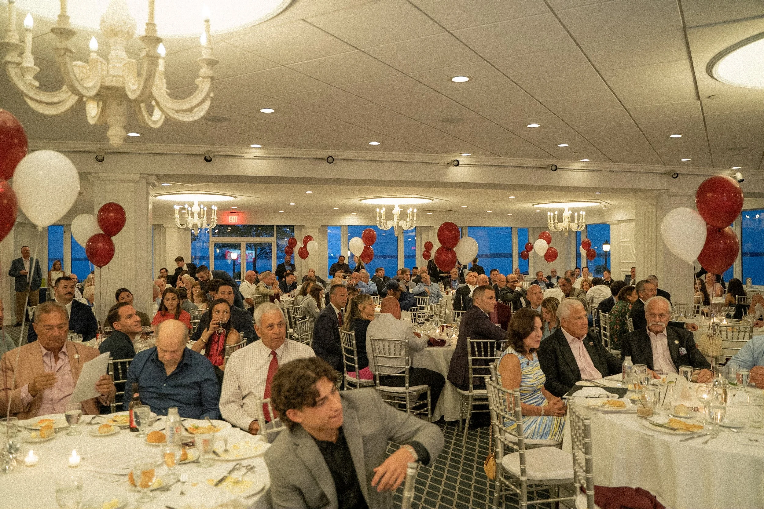 A large banquet hall filled with people attending a celebration or event, decorated with numerous red and white balloons, chandeliers, and large windows showing an evening sky outside.