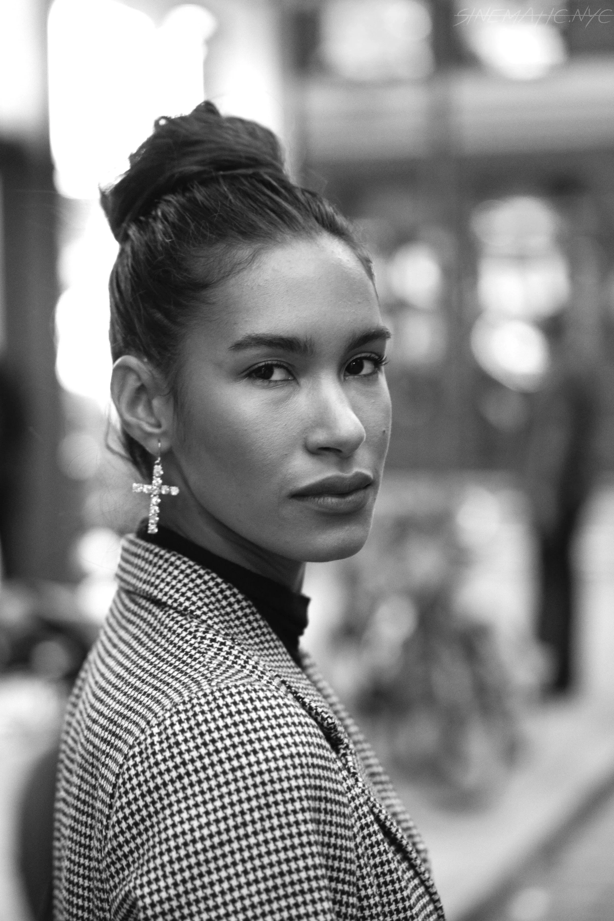 Black and white portrait of a woman with slicked back hair in an updo, wearing cross earrings and a houndstooth patterned blazer, looking over her shoulder with a serious expression, blurred background.