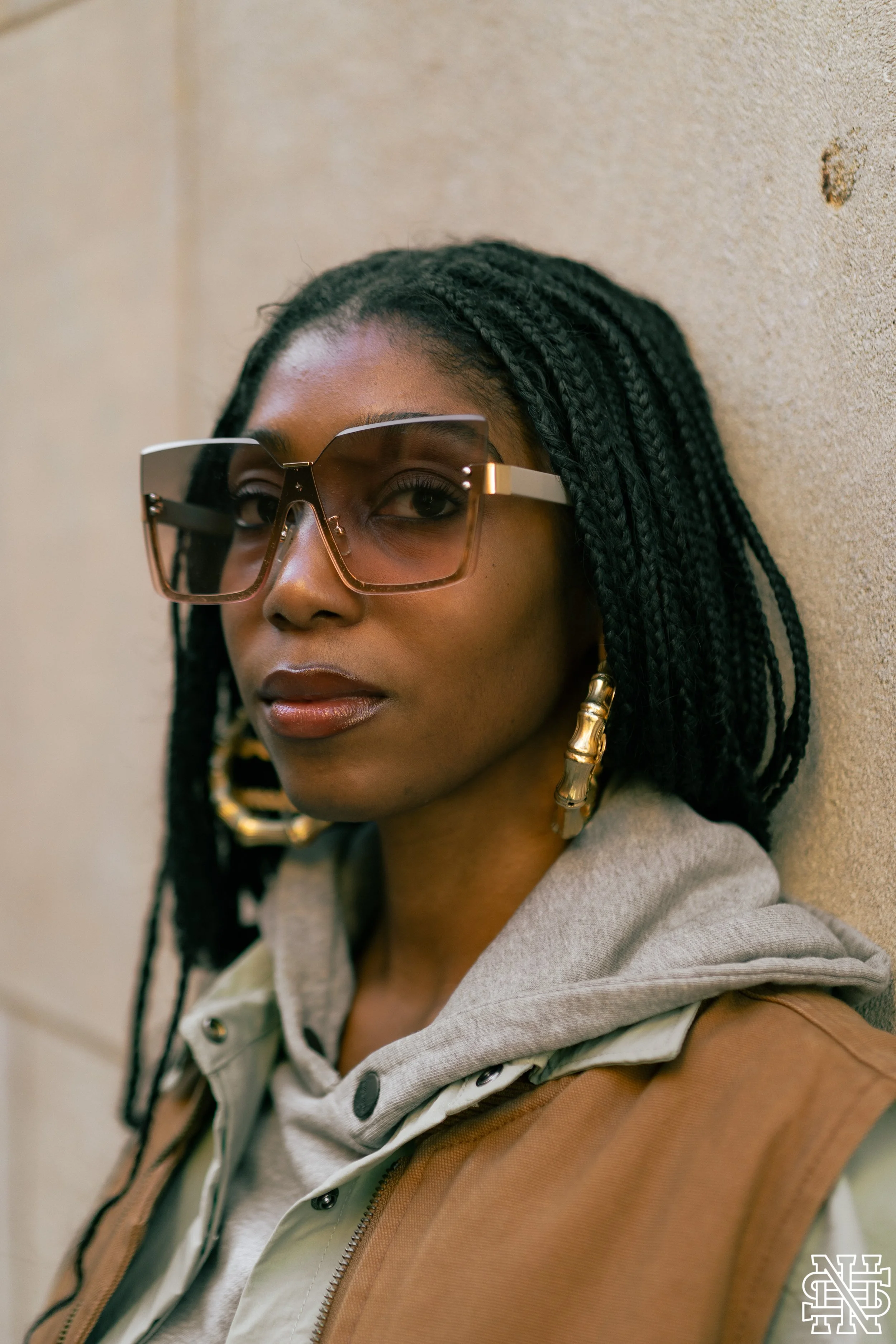 A woman with braided hair, wearing large sunglasses with gold accents, hoop earrings, a gray hoodie, and a brown vest, leaning against a beige wall.