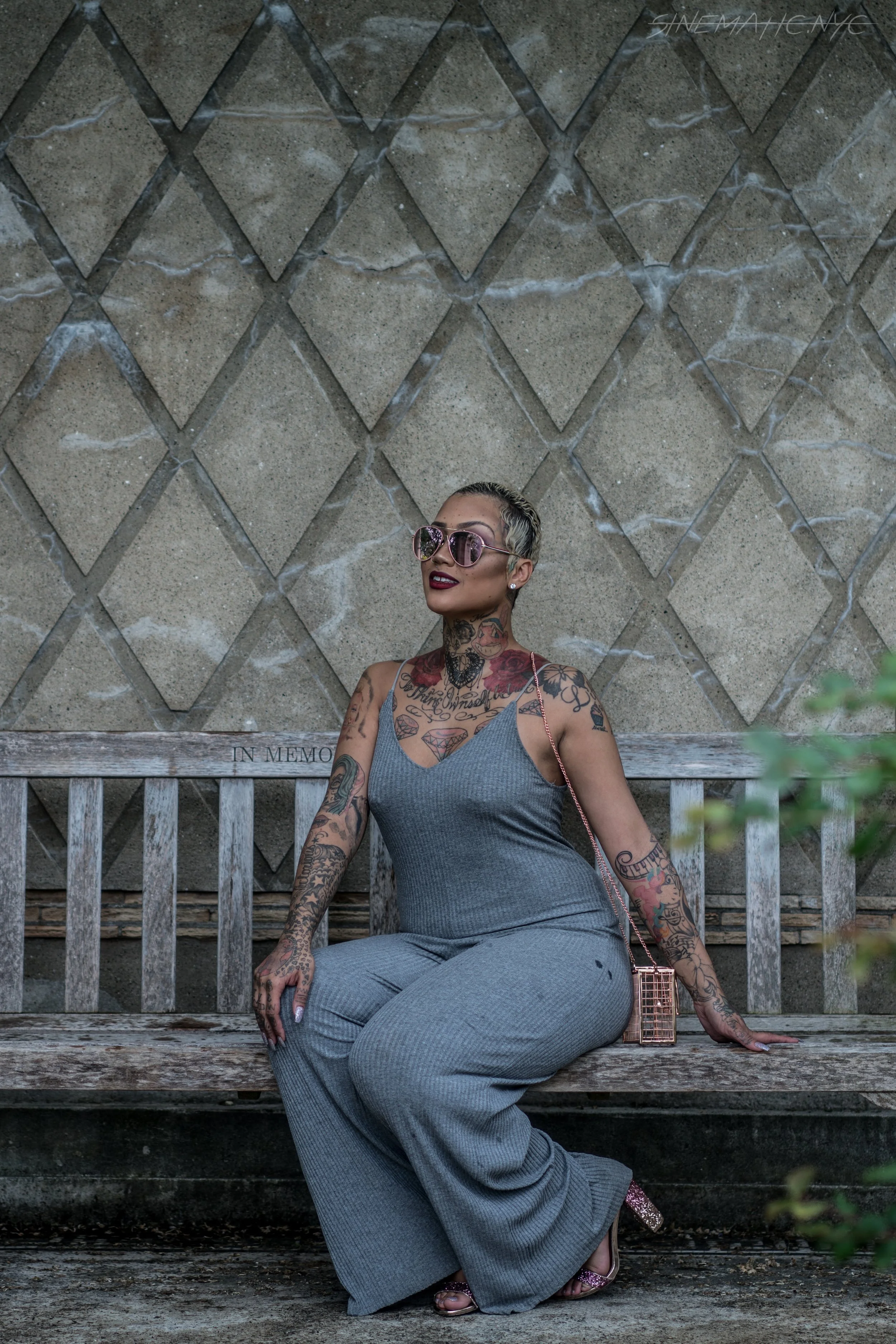 A tattooed woman wearing sunglasses and a gray jumpsuit sitting on a wooden park bench with a stone wall in the background.