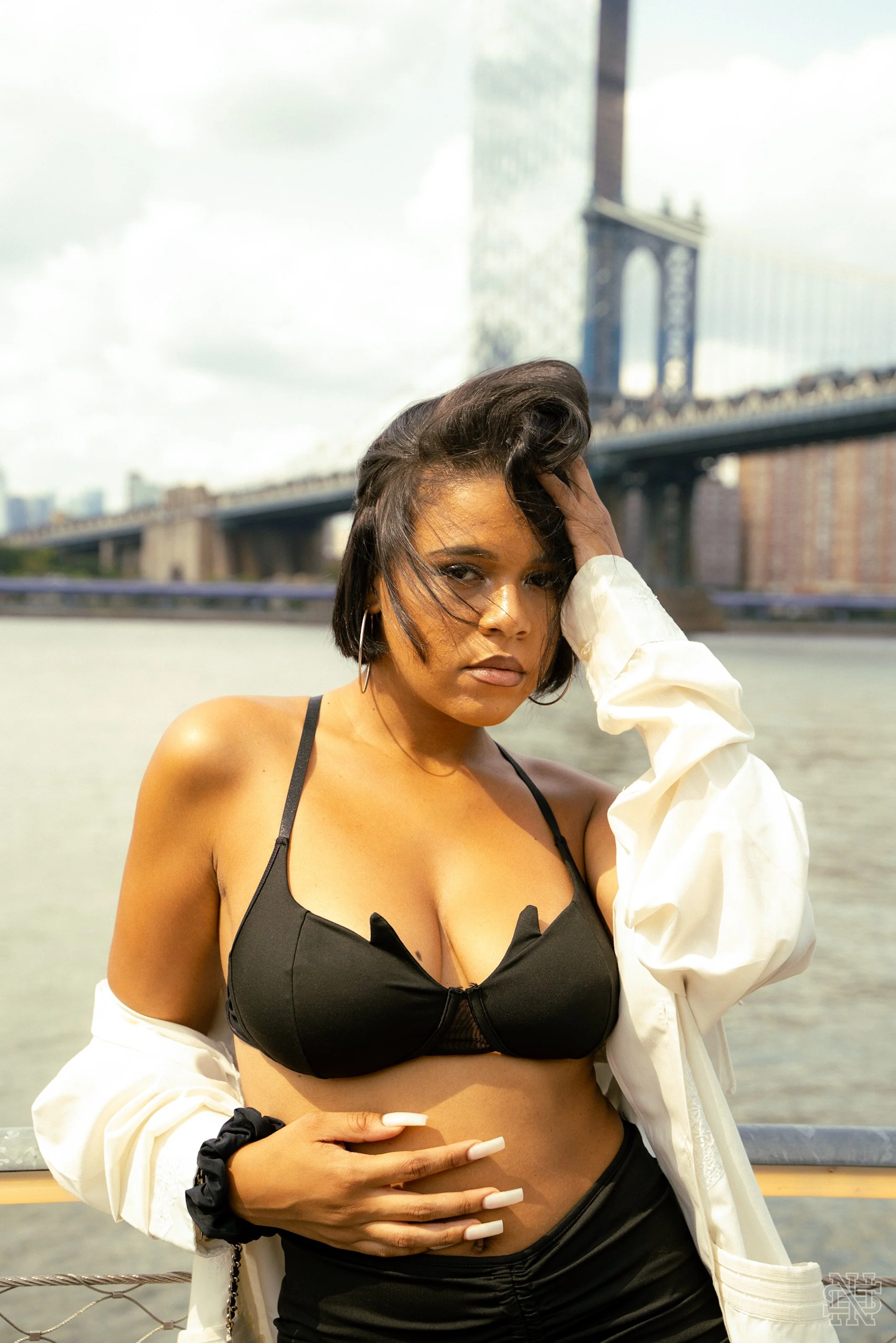 A woman with short dark hair standing in front of a river, wearing a black bra and black pants with white jacket draped over her shoulders, and a bridge and city skyline in the background.