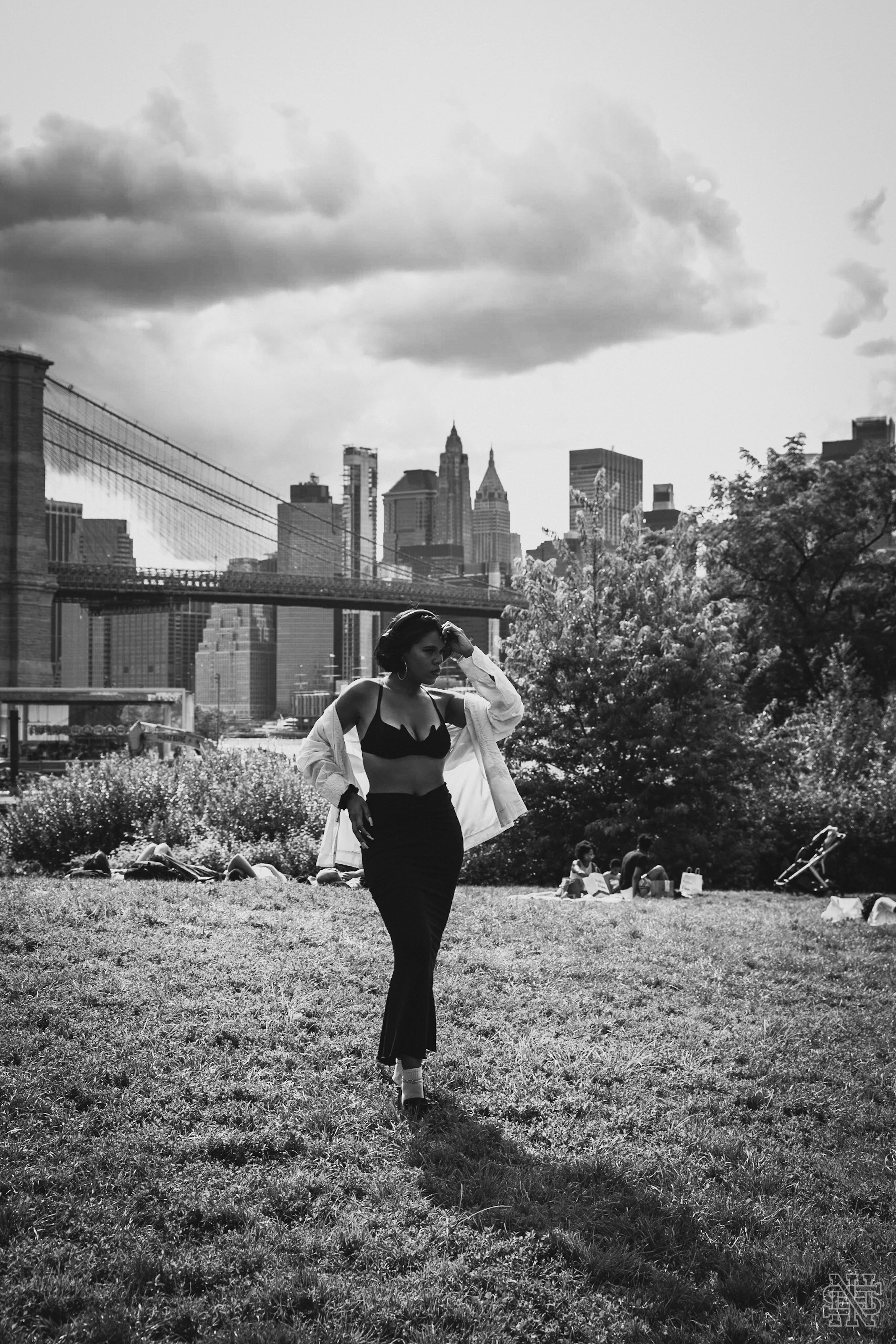 A woman standing on a grassy area in a city park with the Brooklyn Bridge and New York City skyline in the background, dressed in a black bra and high-waisted pants, with a white jacket draped over her shoulders.