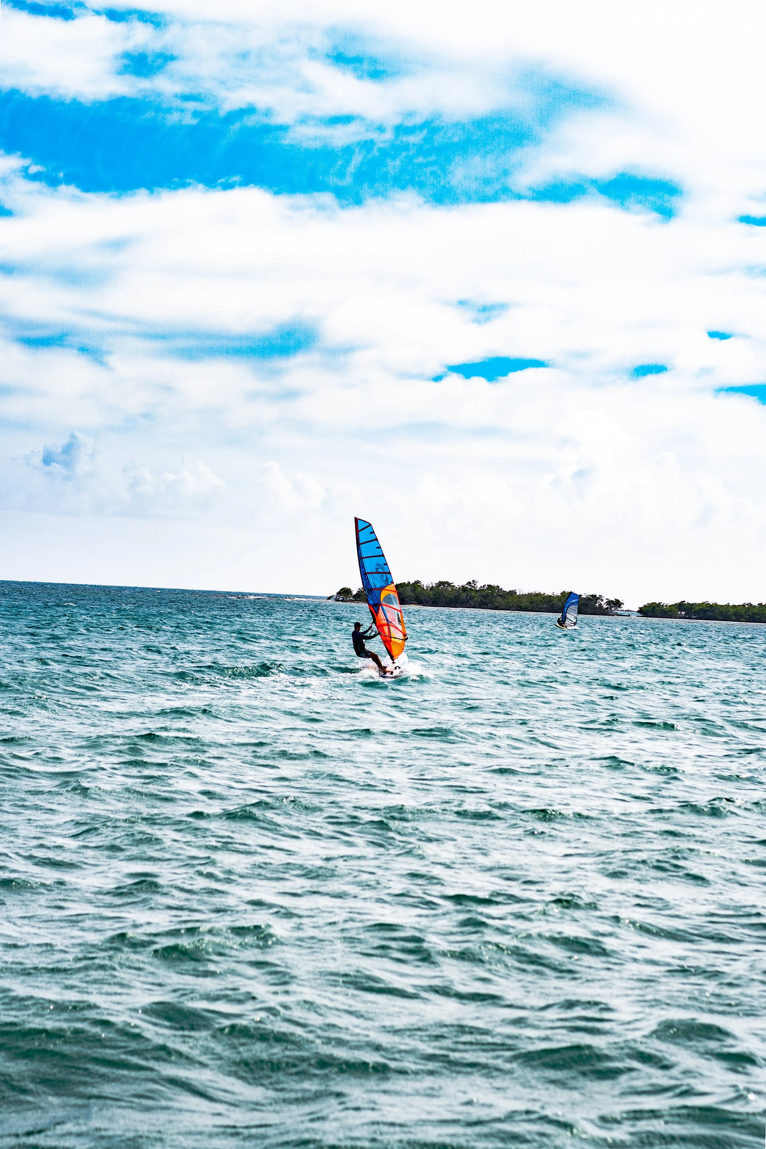 A person windsurfing on the ocean with a colorful sail, and another windsurfer in the background, under a partly cloudy sky.