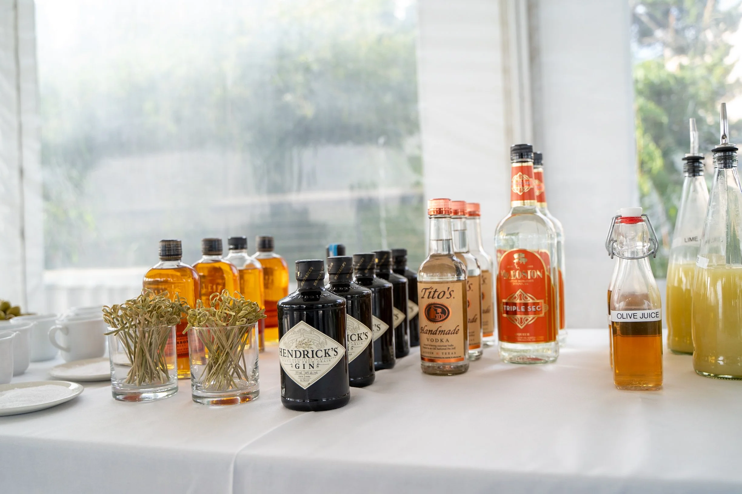 A table with various liquor bottles, small cups, and containers of garnish and juice, set in front of a window with natural light.