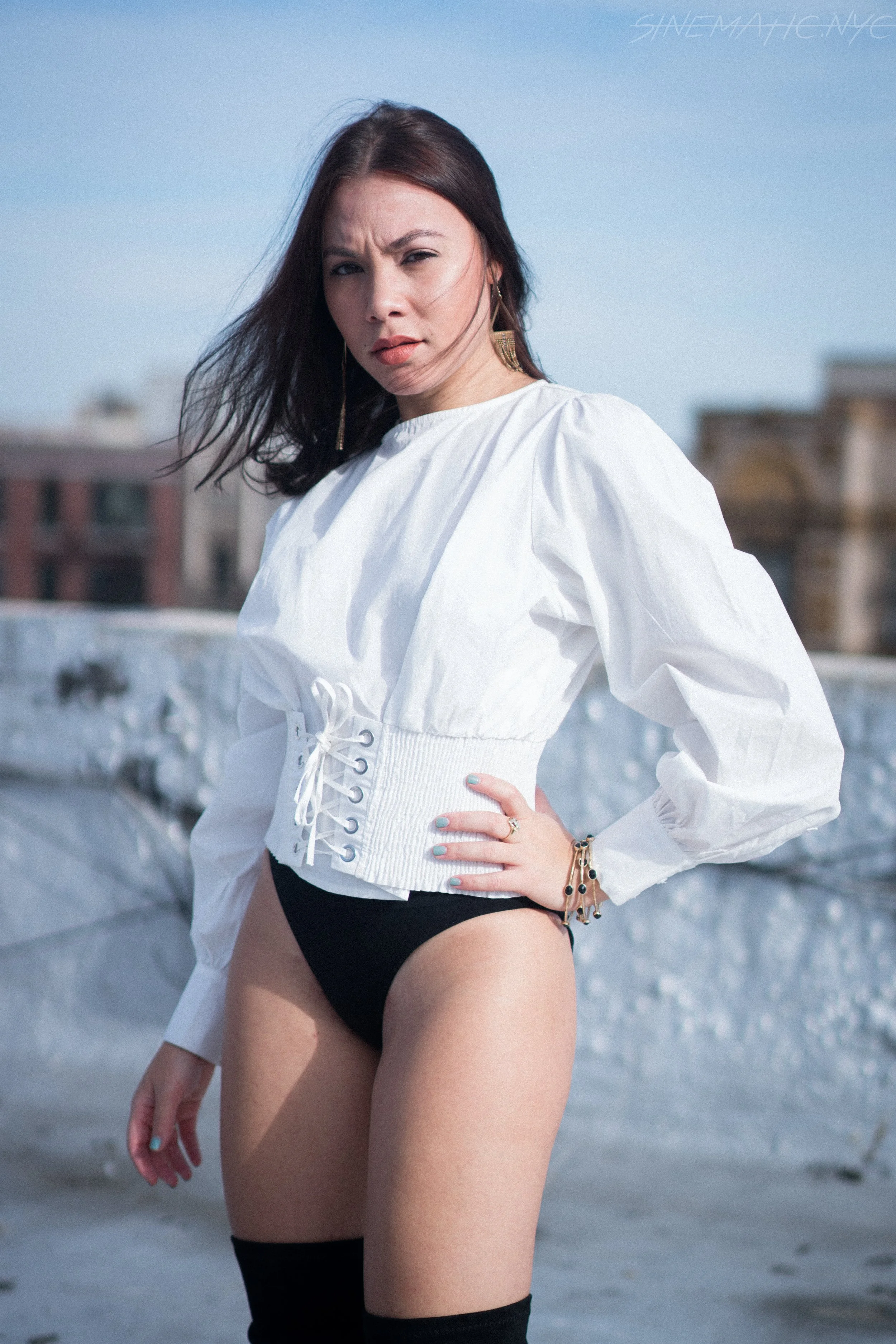 A woman with dark hair standing on a rooftop, wearing a white long-sleeve blouse with puffed sleeves, black high-waisted underwear, and thigh-high stockings, with city buildings in the background.
