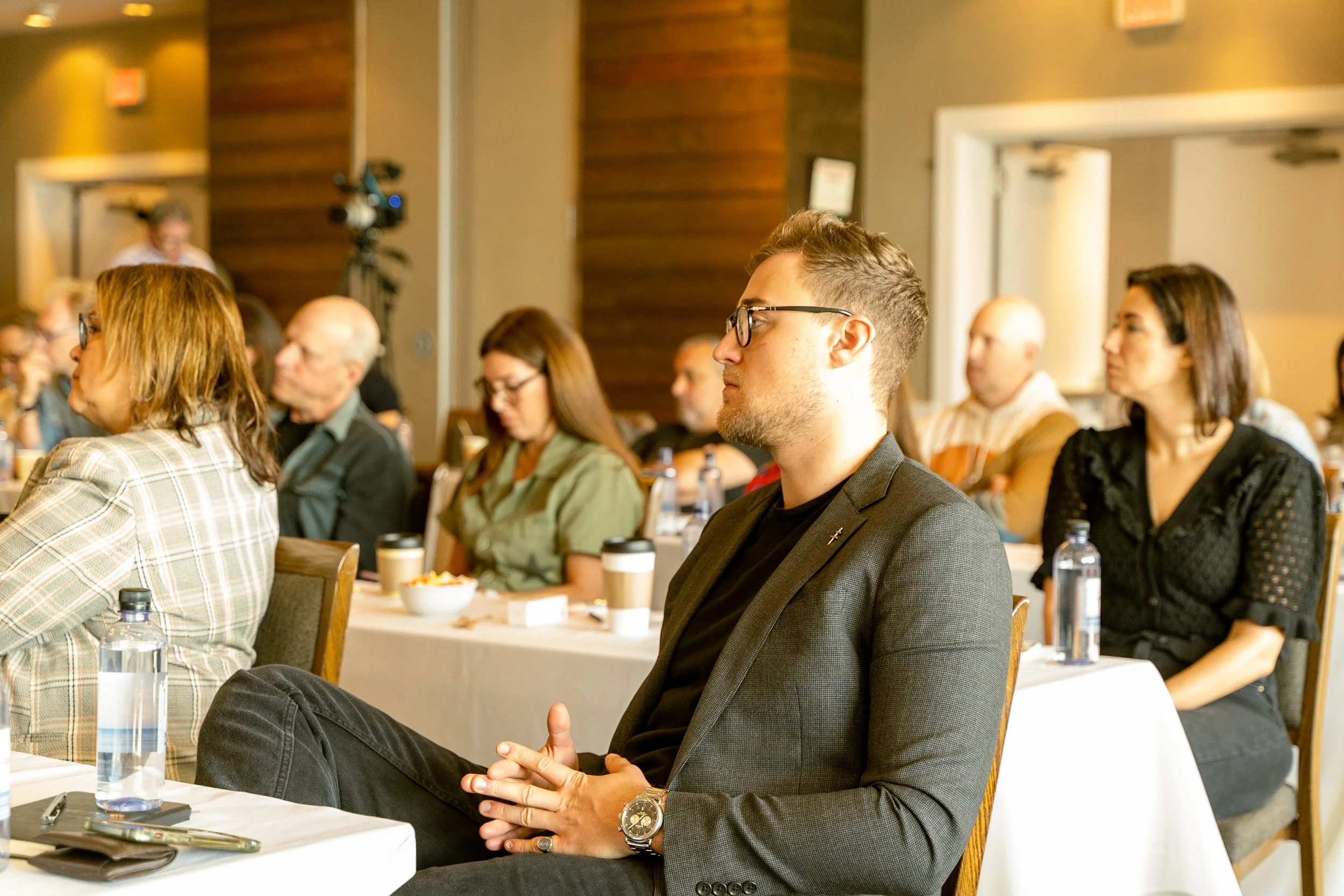 People attending a conference or seminar in a large room, sitting at tables, listening attentively.