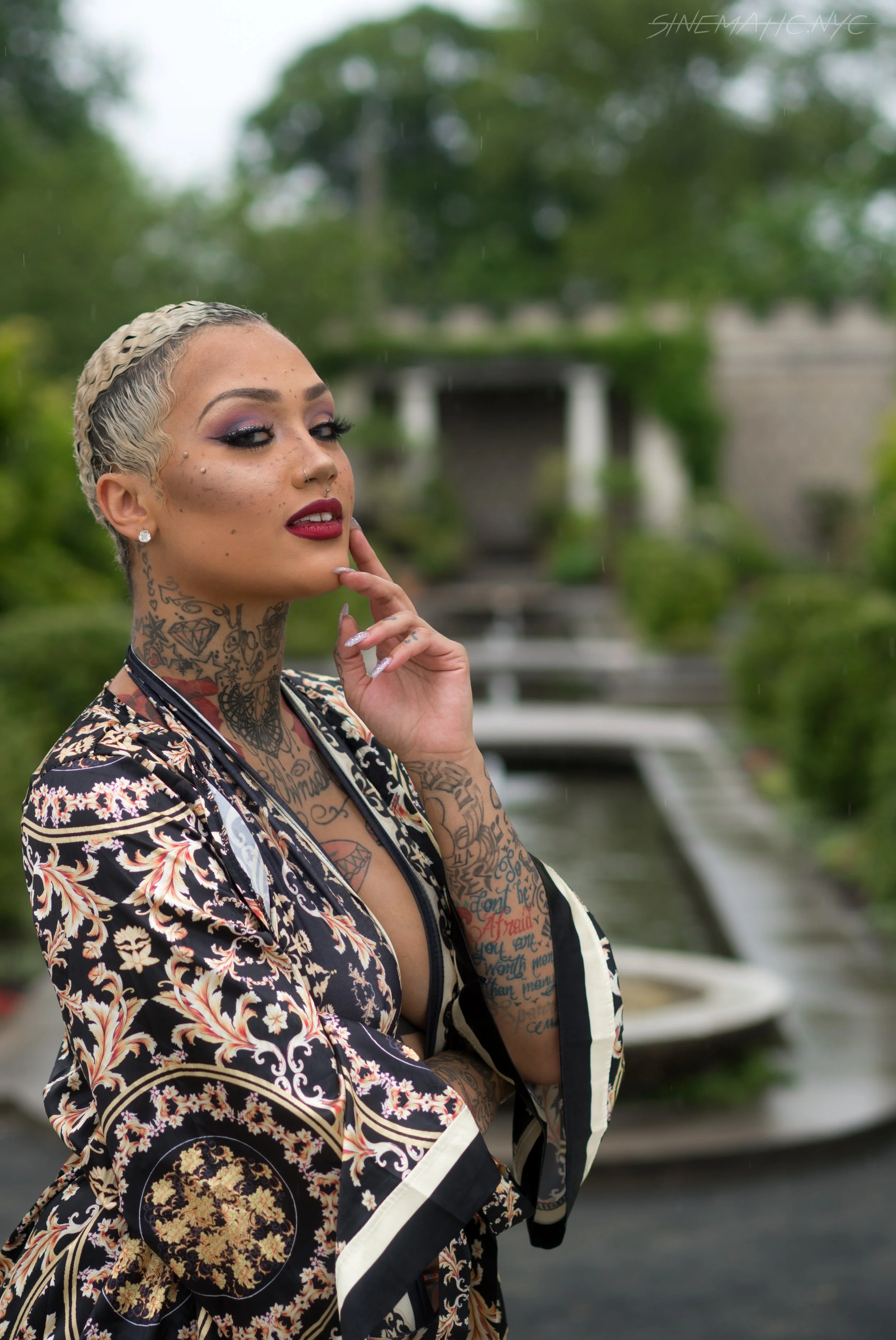 A tattooed woman with short, platinum blonde hair styled in braids, wearing dark eye makeup and red lipstick, posing near a fountain and greenery outdoors.