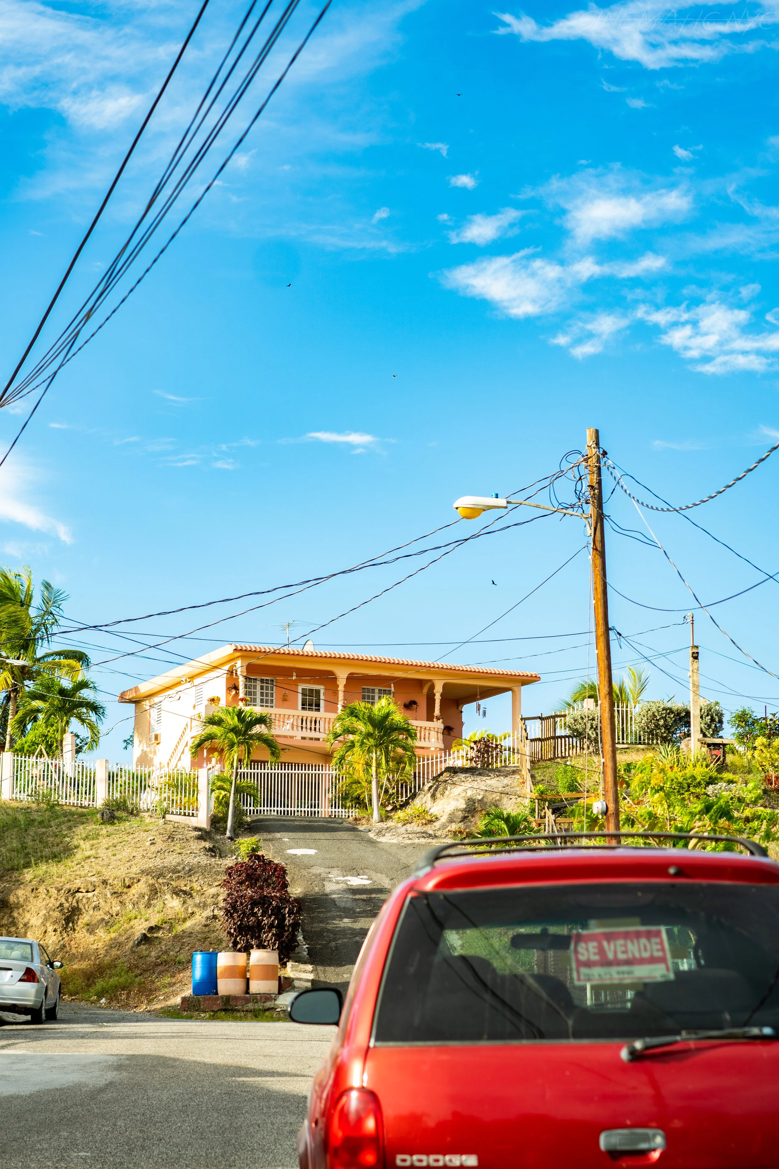 Colorful house on a hill with palm trees, power lines, a blue sky with clouds, and a red car with a 'Se Vende' sign in the window.