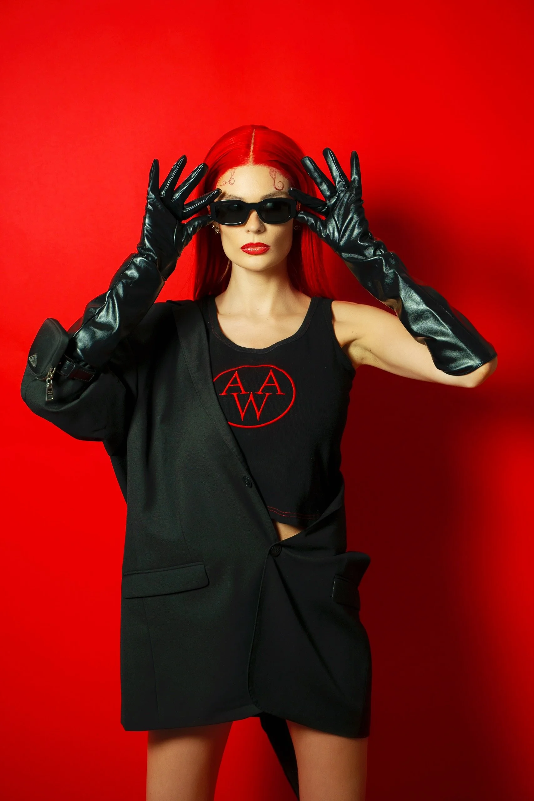 A woman with red hair wearing sunglasses, black gloves, a black tank top with a red logo, and a black blazer against a red background.