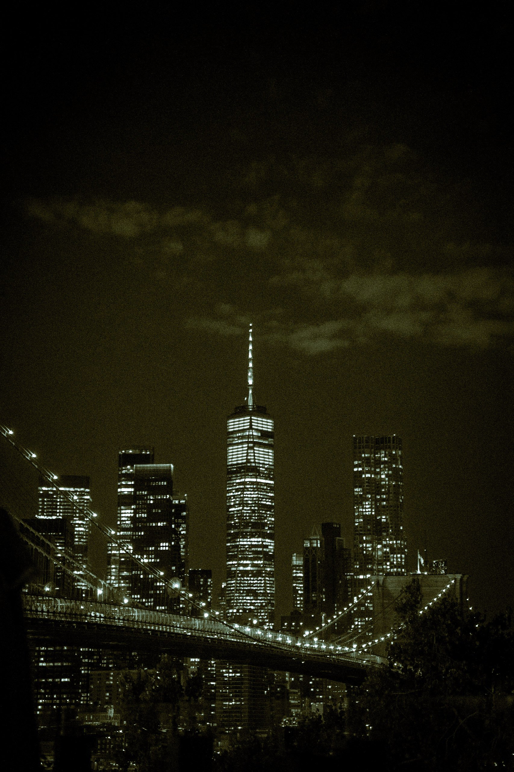 Nighttime cityscape of Manhattan, New York City, featuring the illuminated One World Trade Center building with a tall antenna, surrounded by other skyscrapers and a lit bridge in the foreground.