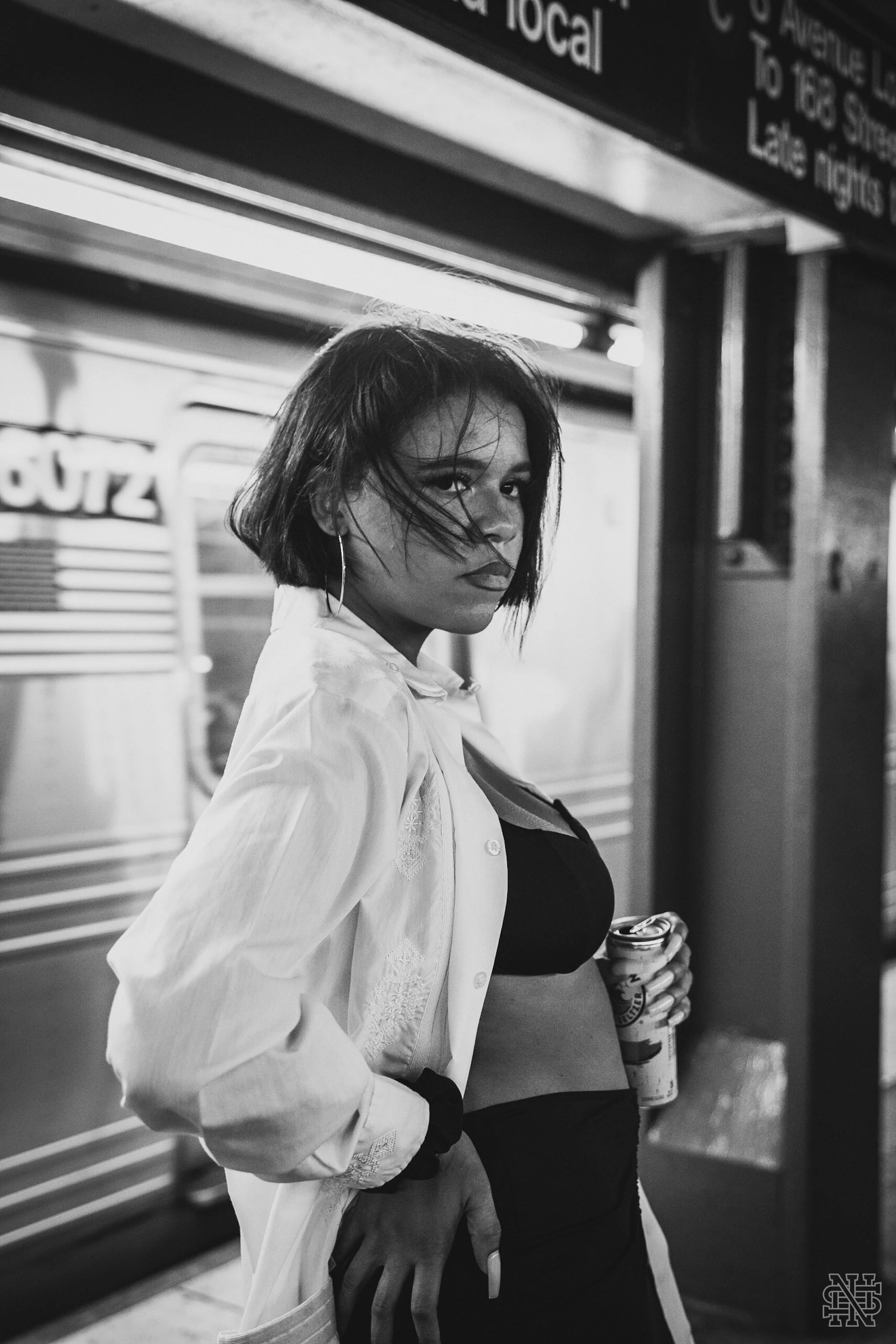 A woman with shoulder-length hair, wearing a white button-up shirt over a black top, stands at a train station holding a drink, looking directly at the camera.