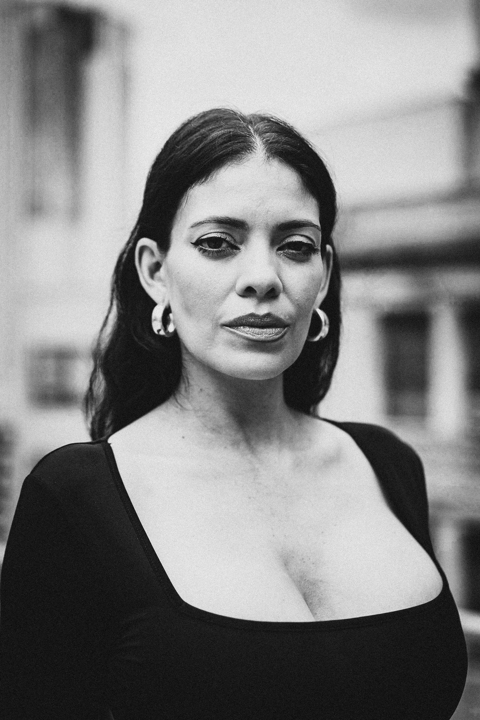 Black and white portrait of a woman with dark hair and bold makeup, wearing a black top with a square neckline and hoop earrings, looking slightly to the side with a confident expression, blurred city background.
