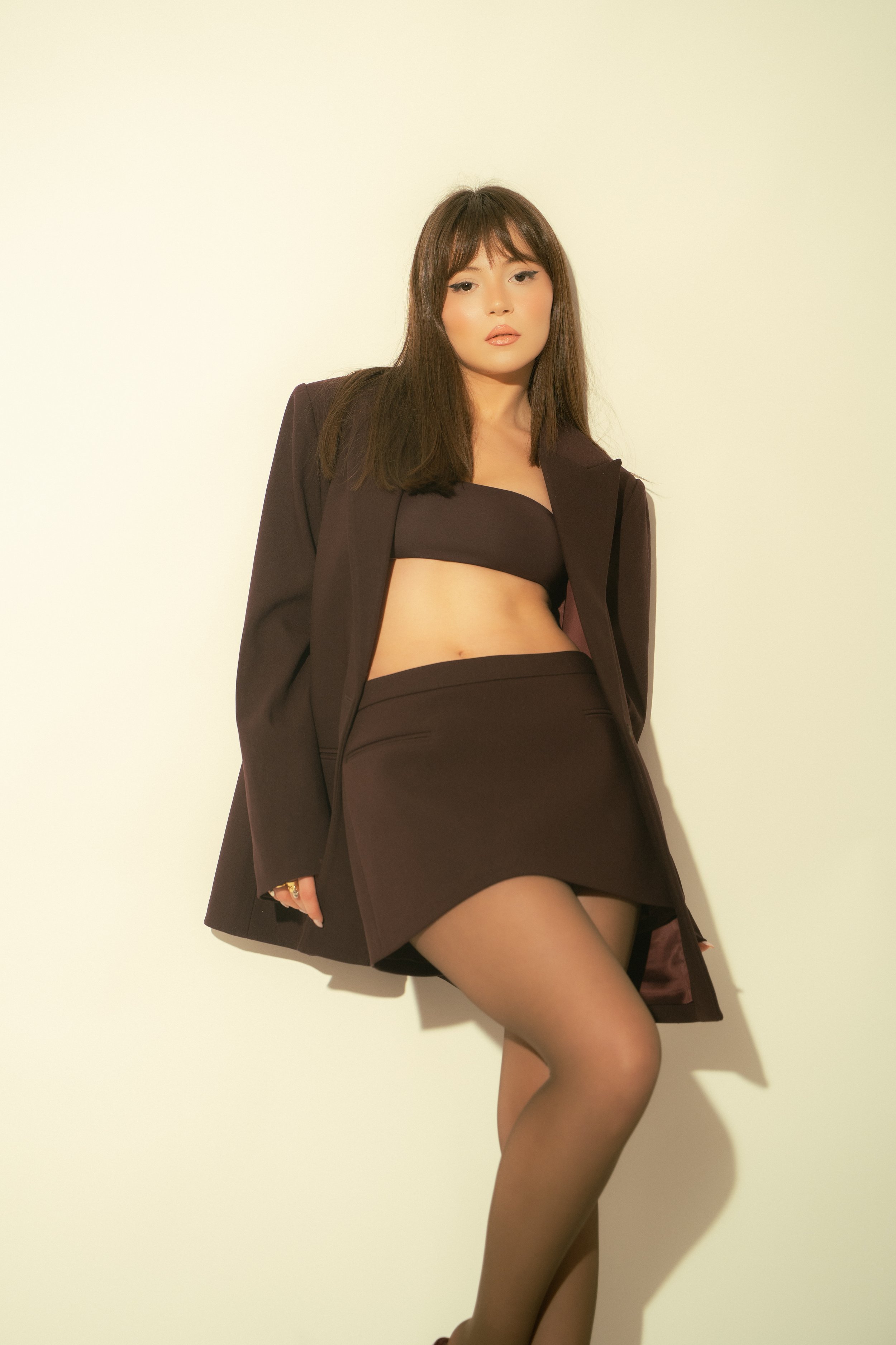 A woman with brown hair wearing a black tube top, black mini skirt, sheer black tights, and a dark blazer stands against a plain light-colored background.