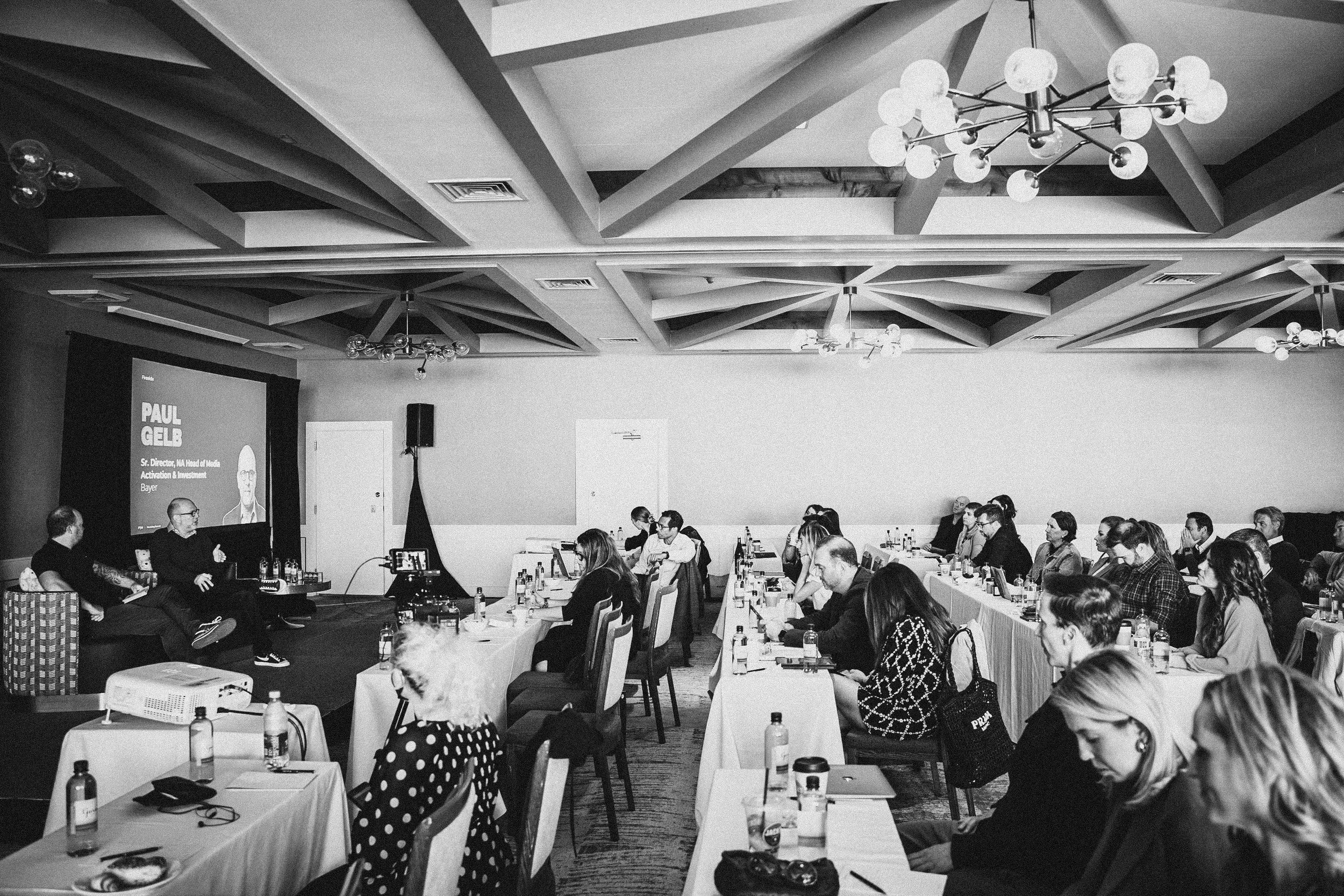 A black and white photo of a conference room filled with seated attendees facing a stage with two speakers. The room has decorative ceiling beams and round ceiling lights, with a large screen displaying a presentation about Paul Gelb.