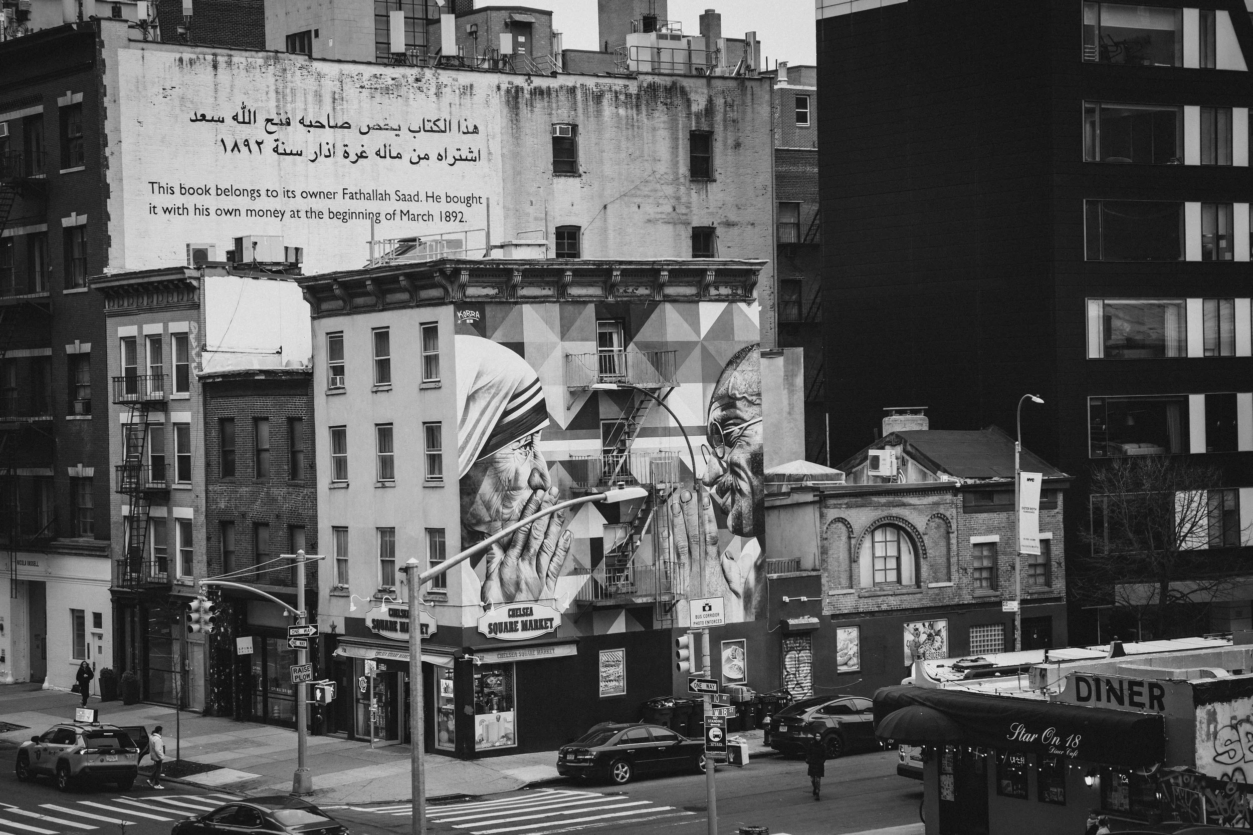 A black and white urban street scene featuring a colorful mural of an elderly woman and a man facing each other. The woman is wearing a headscarf, and the man has glasses. The mural is on the side of a building near Chelsea Square Market, with variou