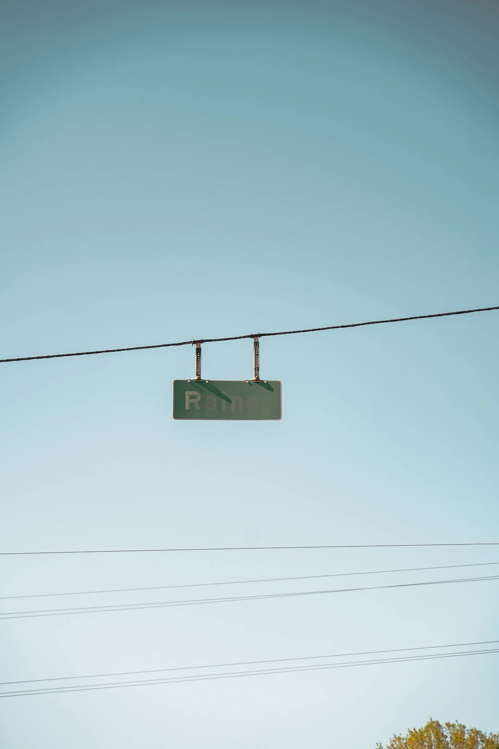 A street sign hanging from a wire against a clear blue sky, with trees visible at the bottom right edge.