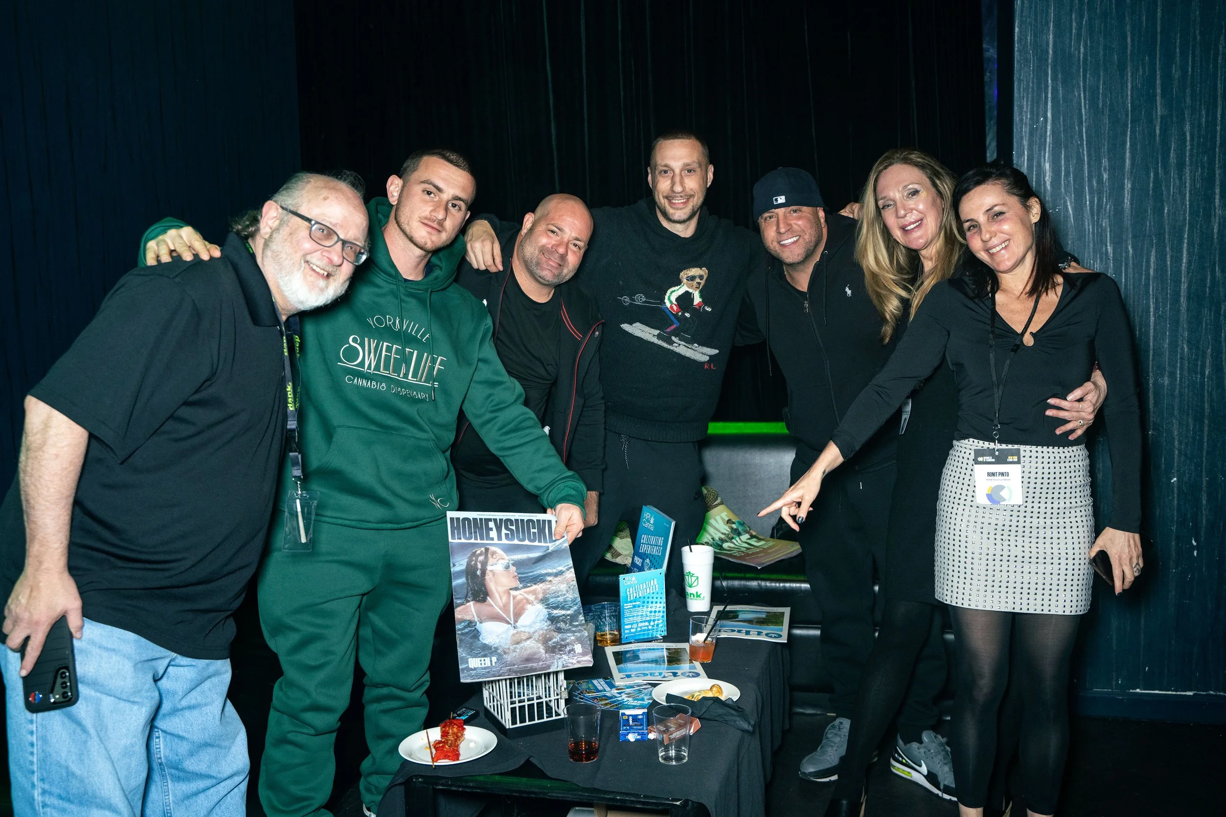 Group of seven people standing together at a social gathering, smiling for the camera, with drinks and magazines on a table in front of them.