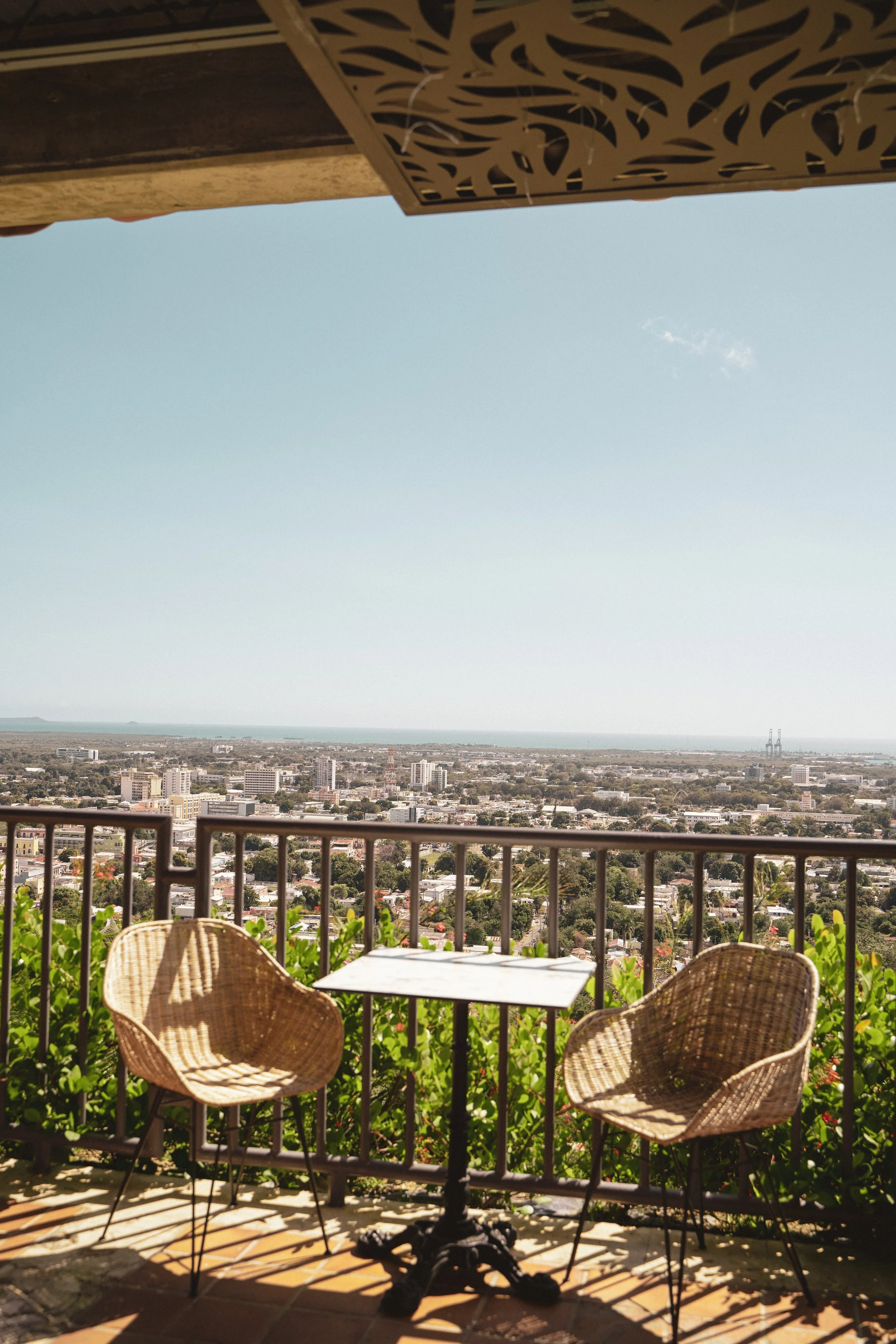 Two wicker chairs and a small table on a balcony overlooking a cityscape with buildings, greenery, and a distant view of water and sky.