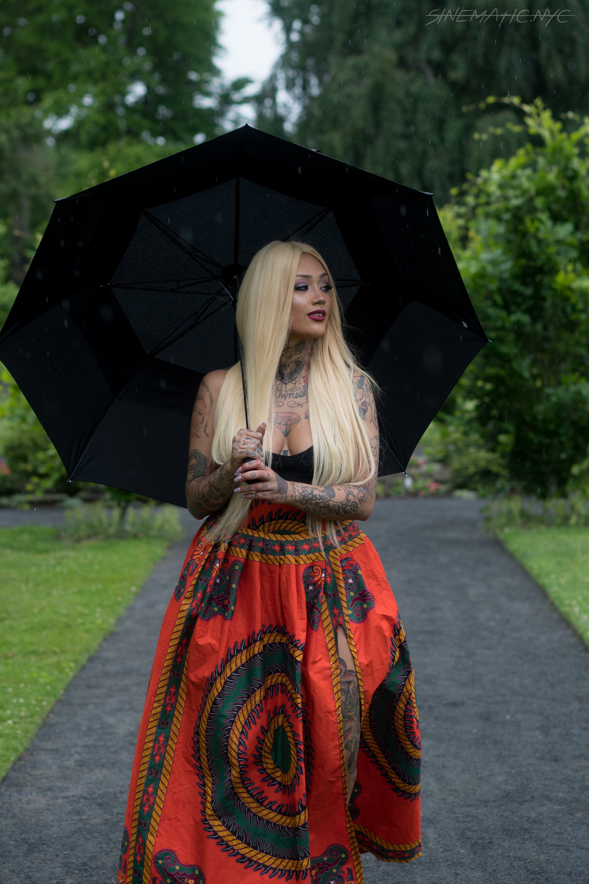 A woman with long blonde hair and tattoos holds a black umbrella standing on a stone path in a park with green trees, rain visible, wearing a black top and a colorful patterned skirt.