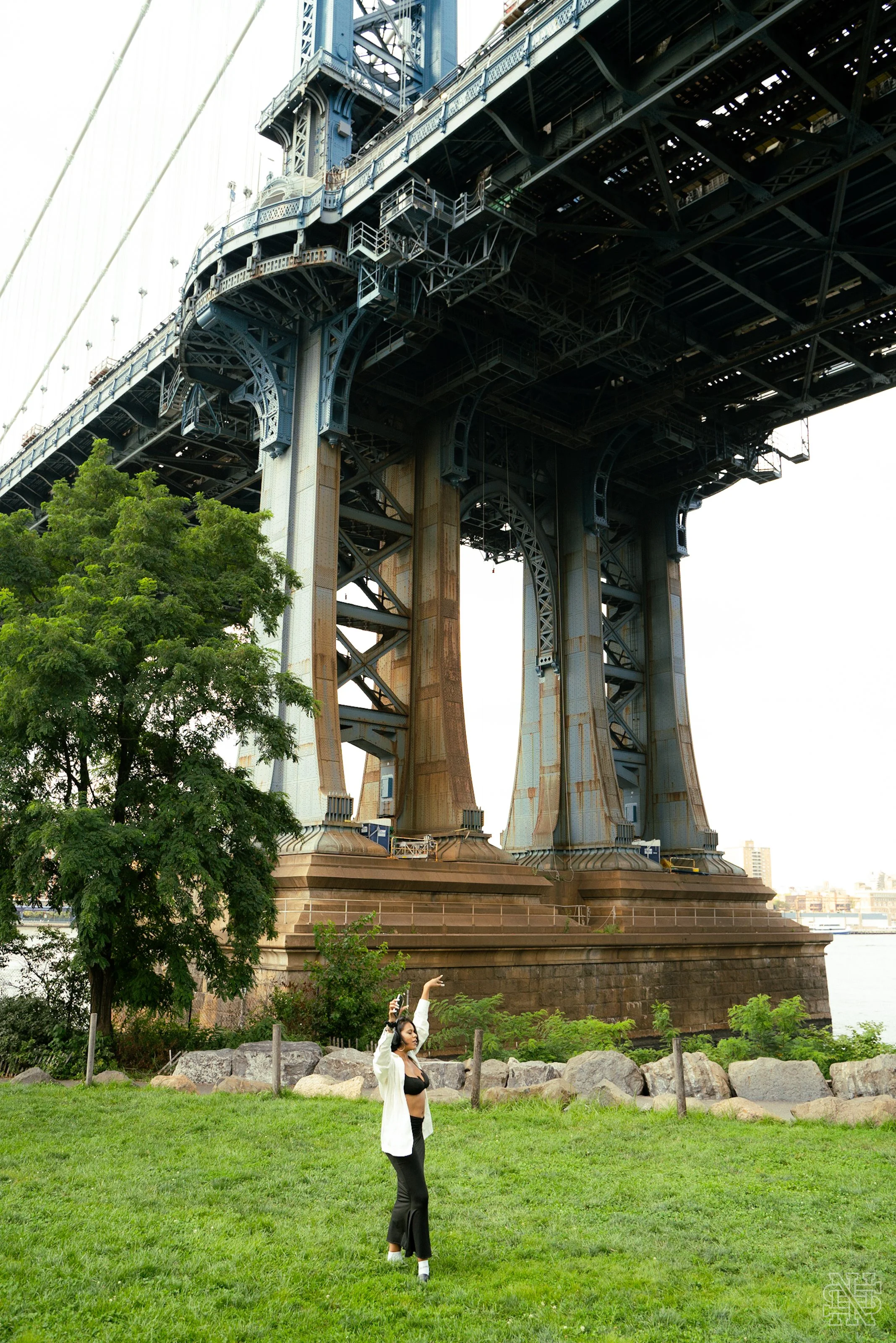 A woman standing on a grassy area under a large suspension bridge, taking a selfie with her phone, with trees and rocks in the background.