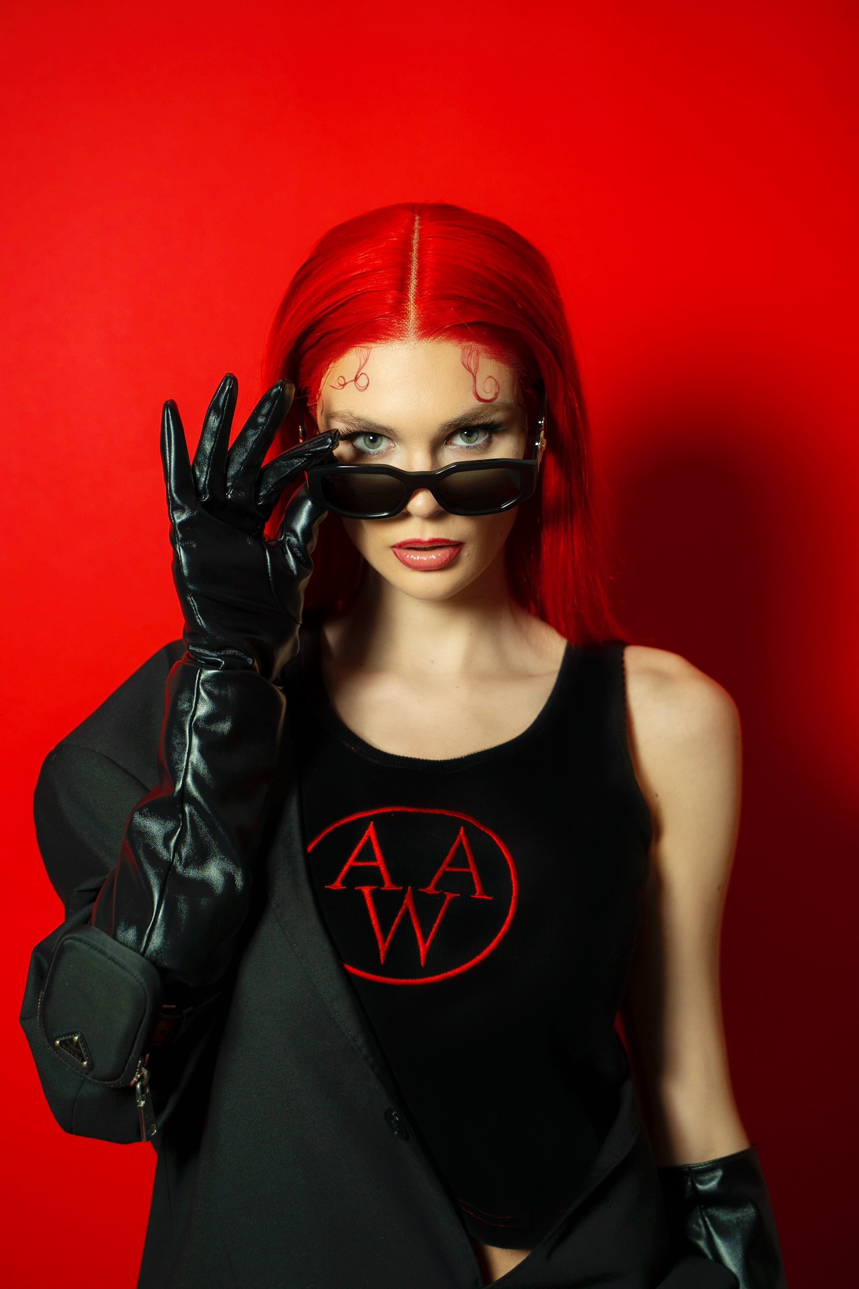 A woman with long red hair, wearing black sunglasses, black gloves, a black jacket, and a black tank top with red lettering, standing against a red background.