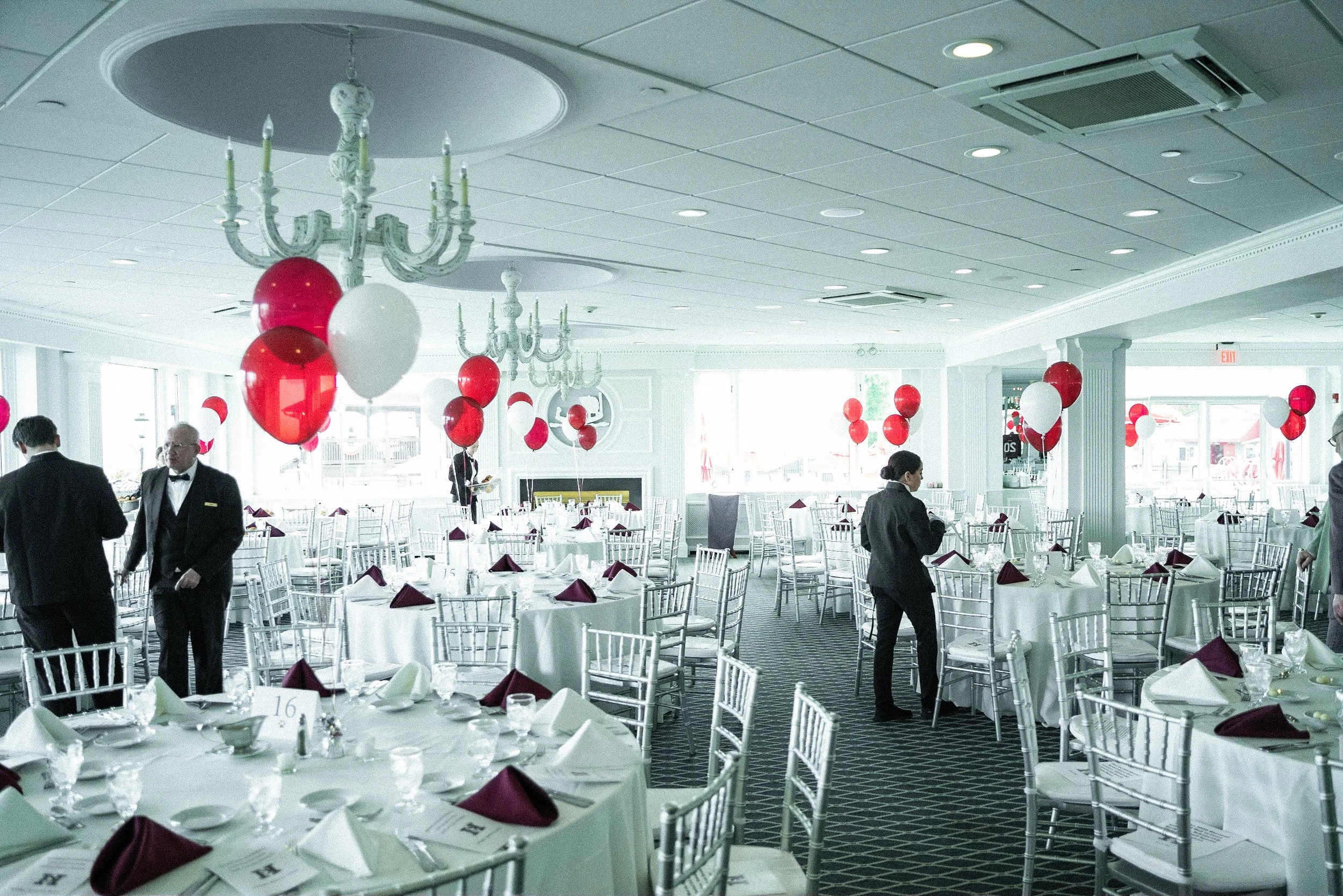 A banquet hall decorated for a celebration with red and white balloons, set with round tables covered in white tablecloths, neatly folded napkins, glassware, and silverware, with a few staff members preparing for the event.