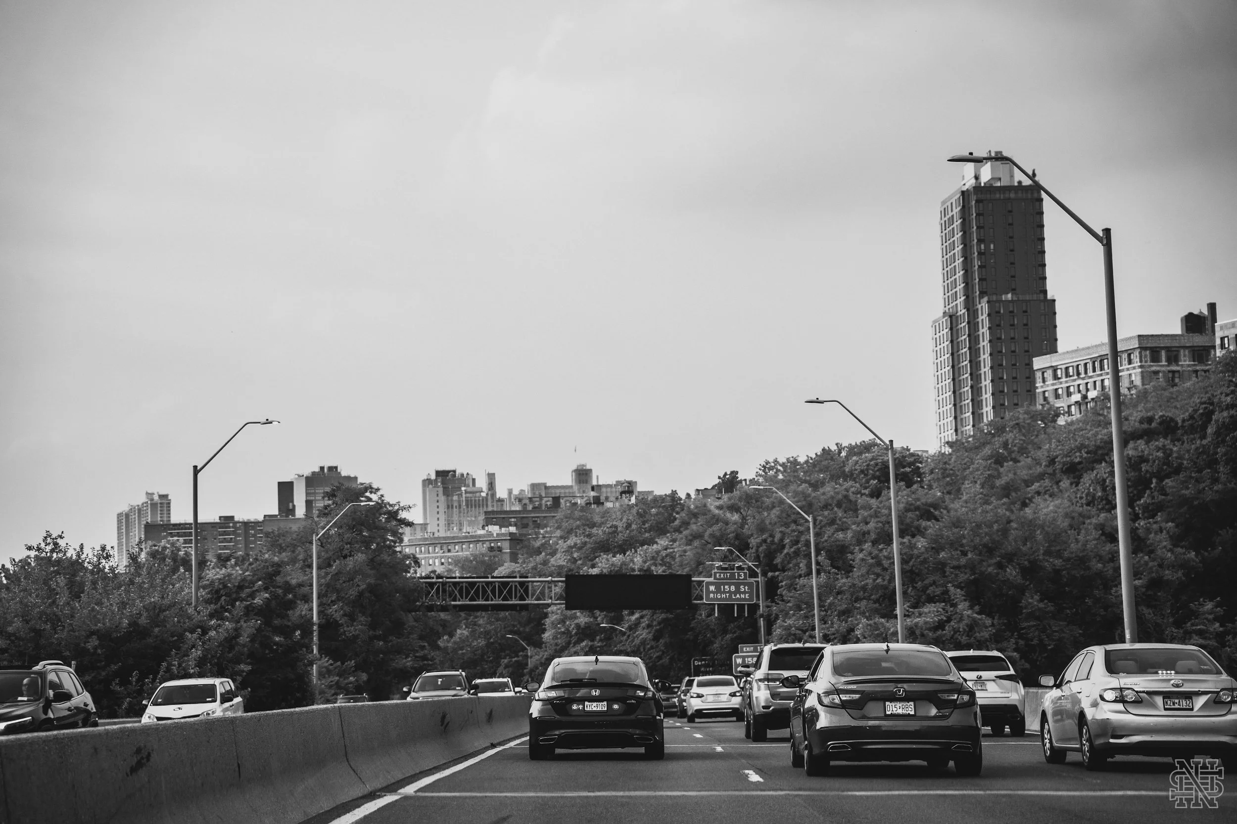 Black and white photo of a busy city highway with multiple cars traveling in both directions, tall buildings and trees in the background, and streetlights along the road.