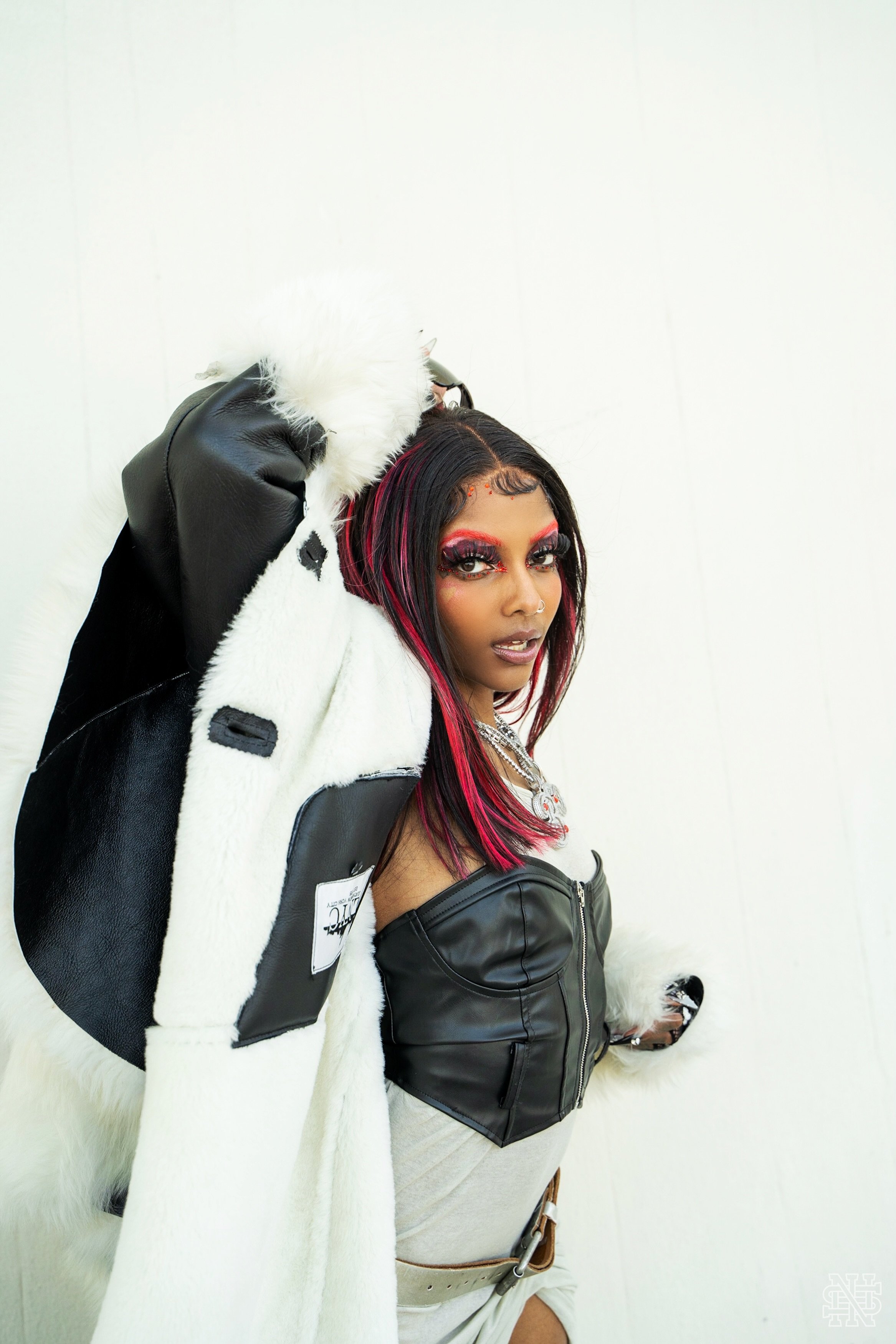 Woman with black and pink hair, dramatic makeup, wearing a black leather top, silver jewelry, and a cream-colored skirt, leaning against a white wall, holding a coat with black leather and white furry elements over her shoulder.