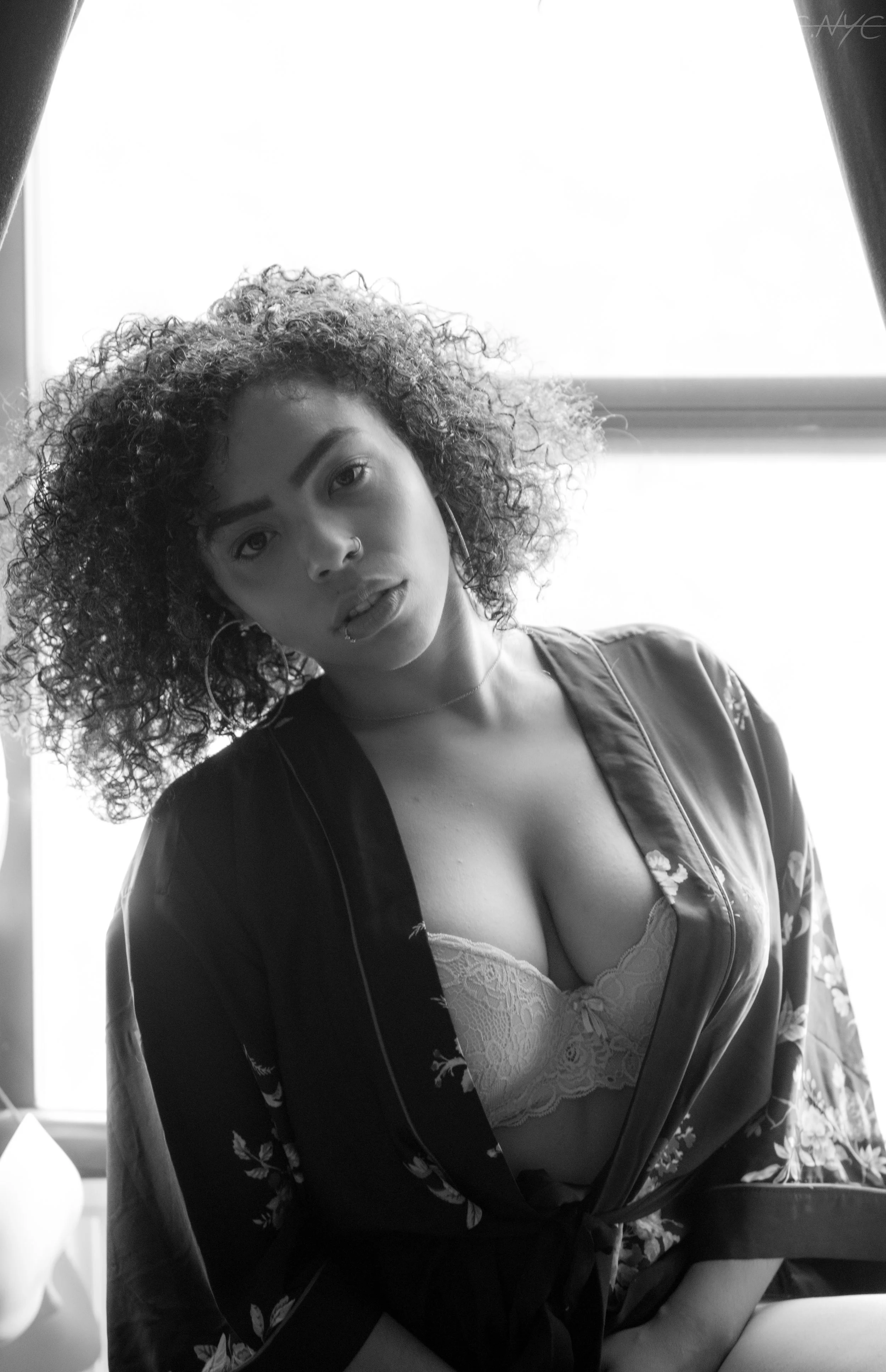 A woman with curly hair, wearing a floral silk robe and lace bra, looking at the camera with a slightly open mouth, standing near a window.