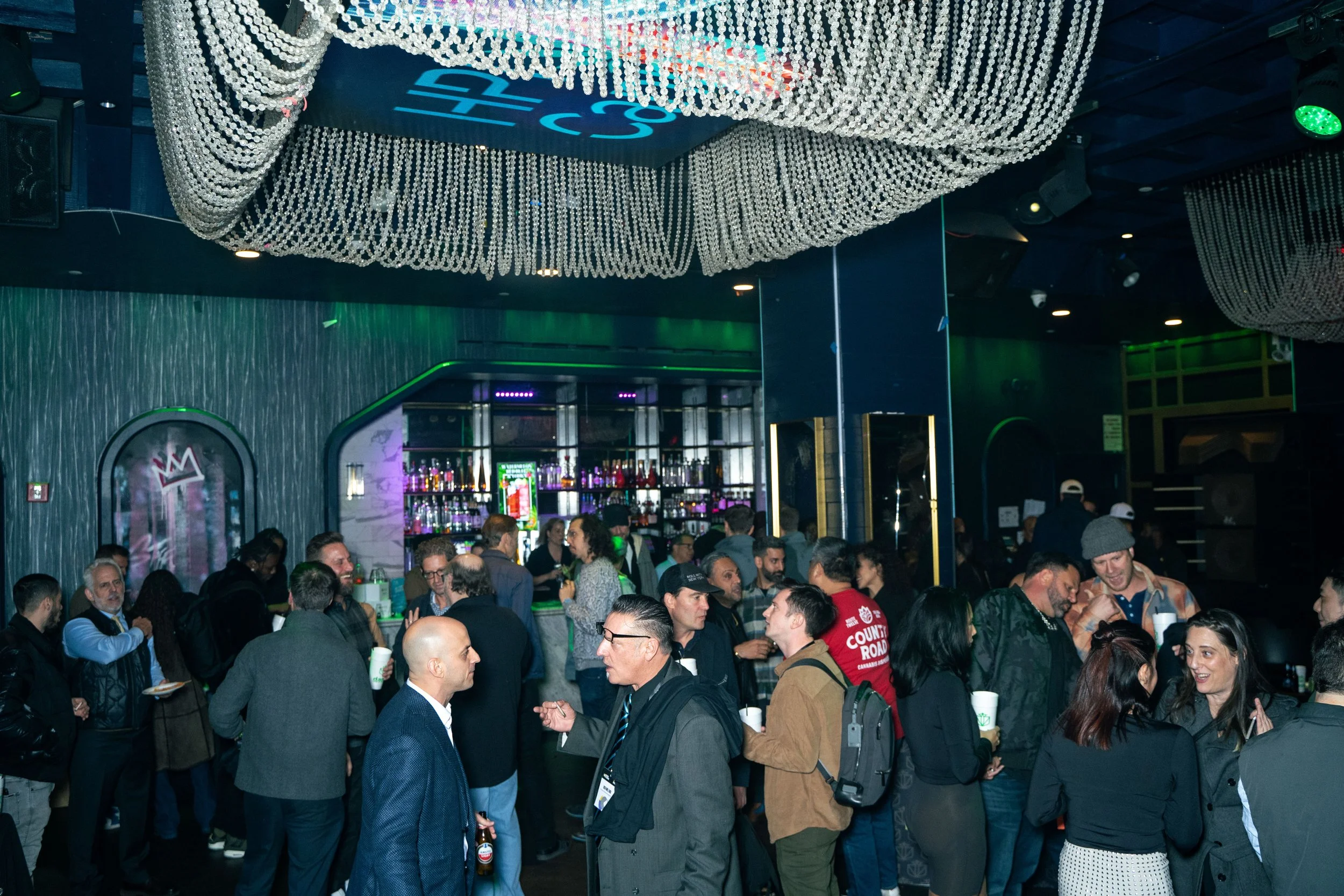 Crowd of people socializing at a crowded indoor bar or nightclub with a decorated ceiling, bar shelves with bottles, and colorful lighting.