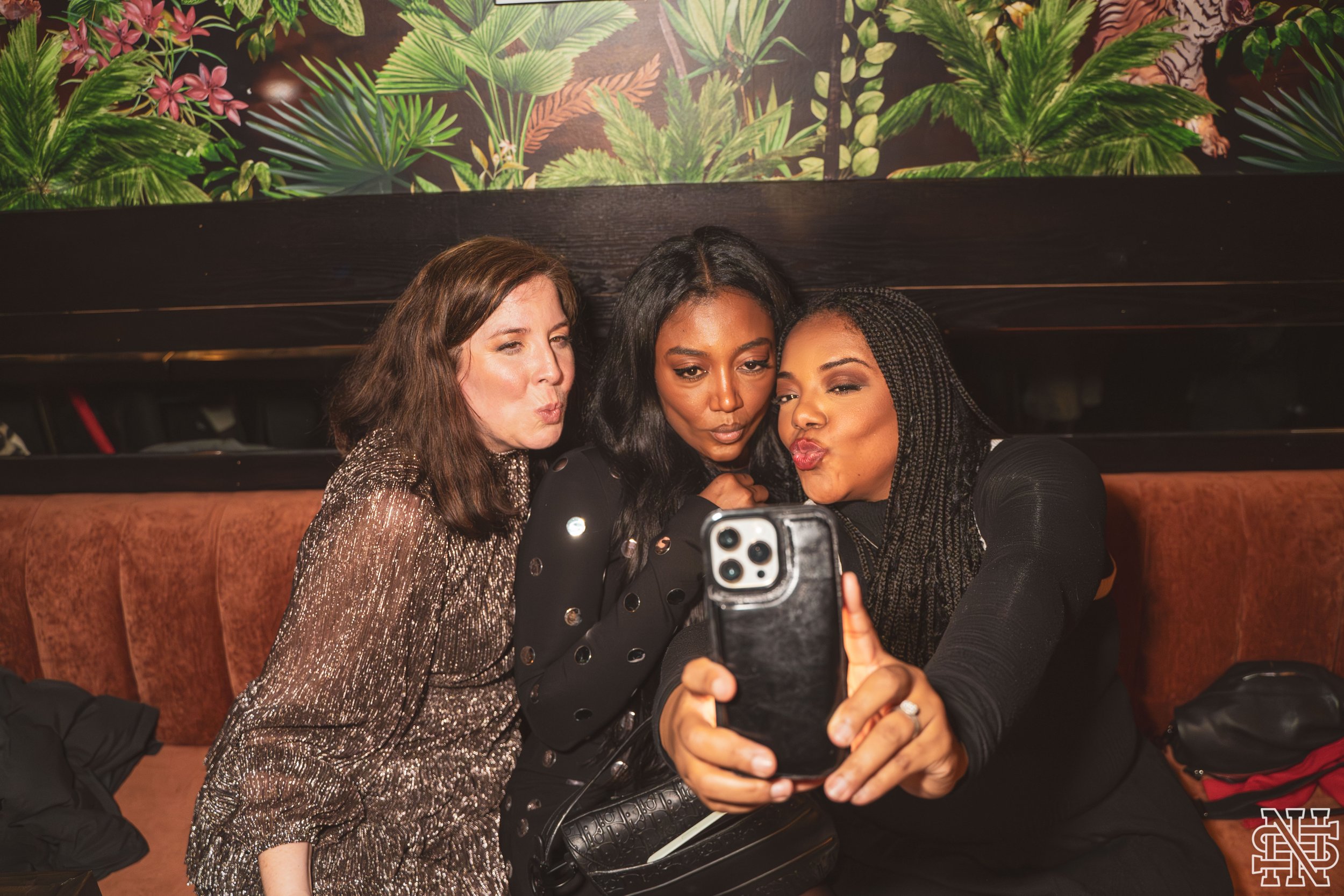 Three women sitting on a brown leather couch taking a selfie together, with a tropical-themed wallpaper background.