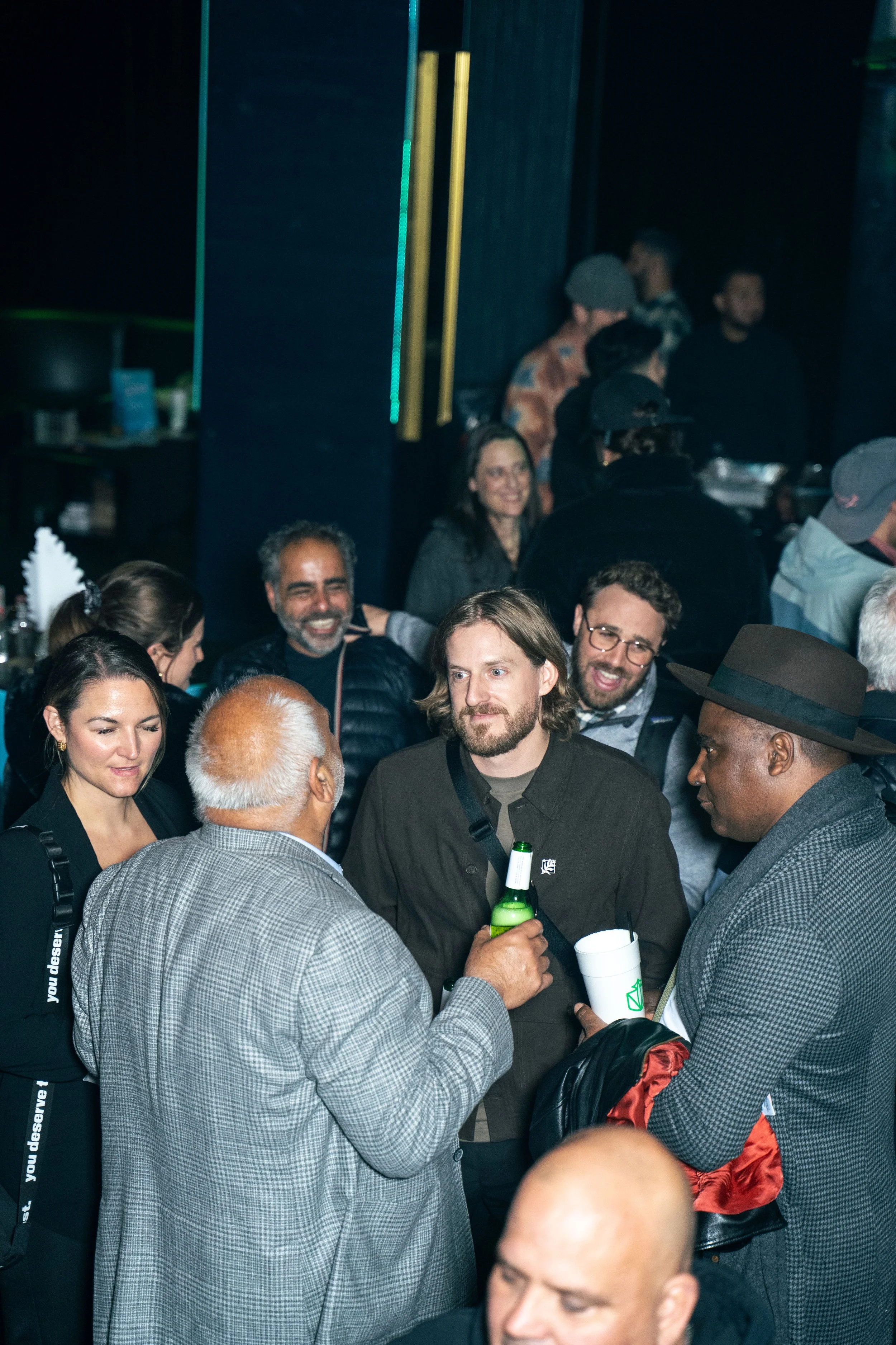Group of diverse people socializing at an indoor event, some holding drinks, engaged in conversation, with a dark background and some neon lights.