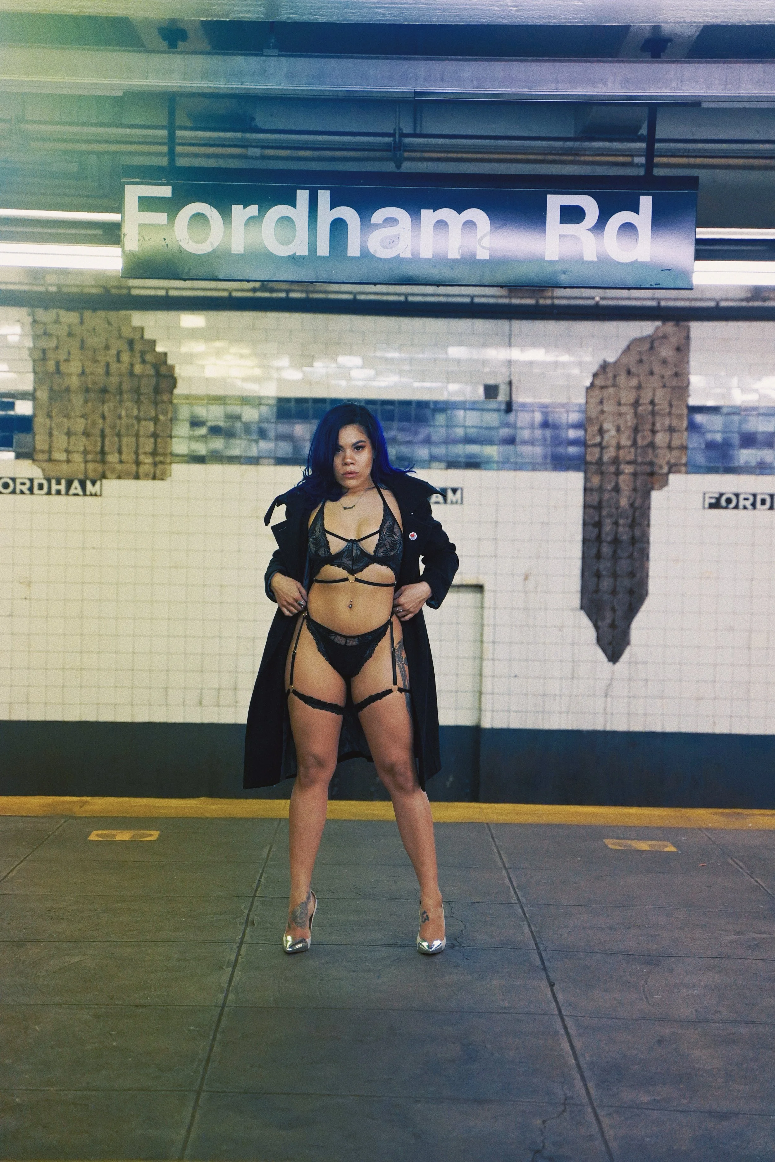 A woman standing on a subway platform at Fordham Road station, wearing revealing black lingerie and a long black coat with tattoos on her legs and arms.