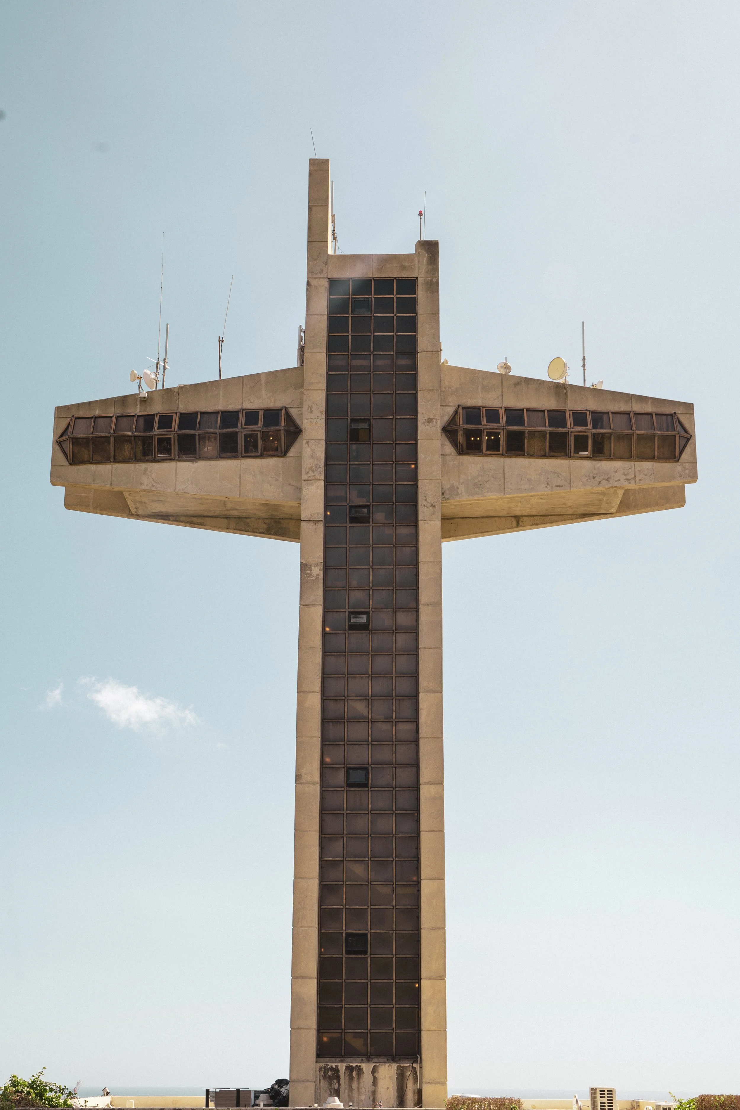 A tall, concrete building shaped like a cross, with windows on the arms and a vertical section in the middle, against a clear blue sky.