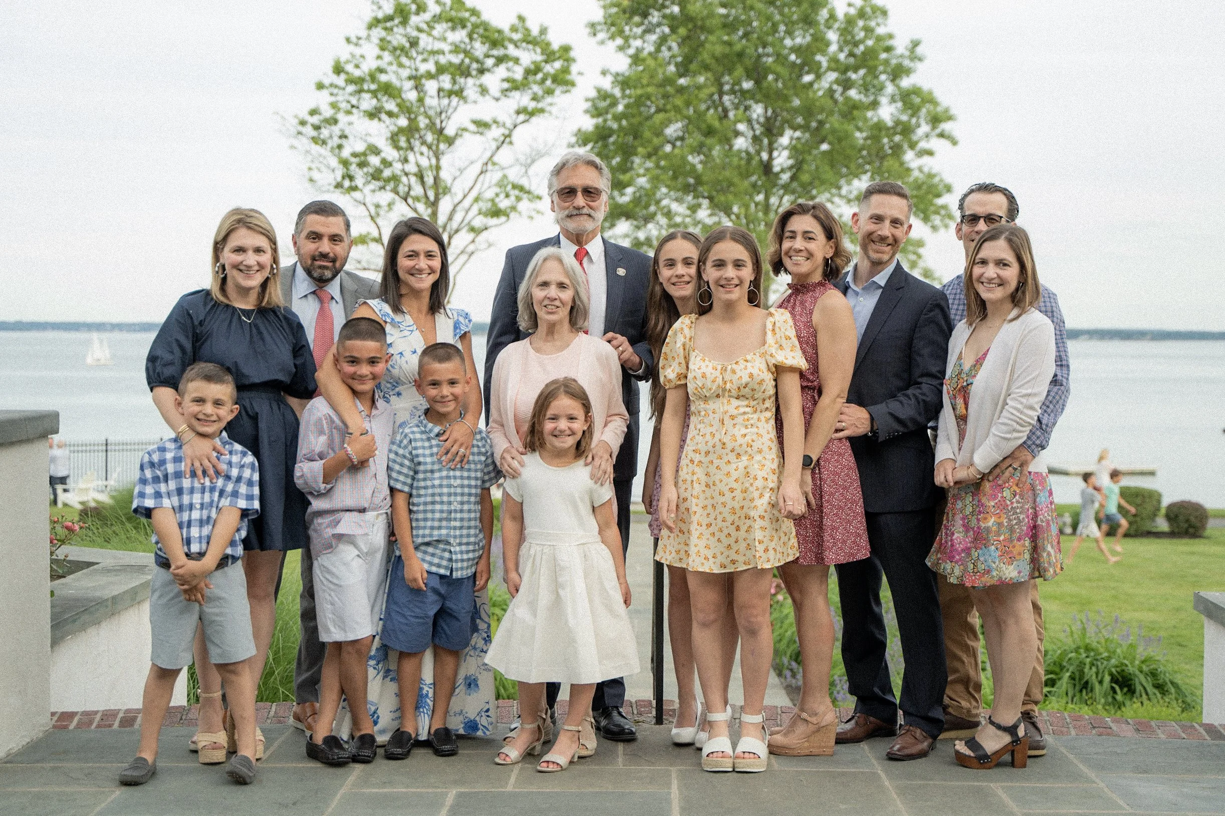 Group of 15 people, including children and adults, posing outdoors near water with a sailboat, green trees, and children playing in the background.