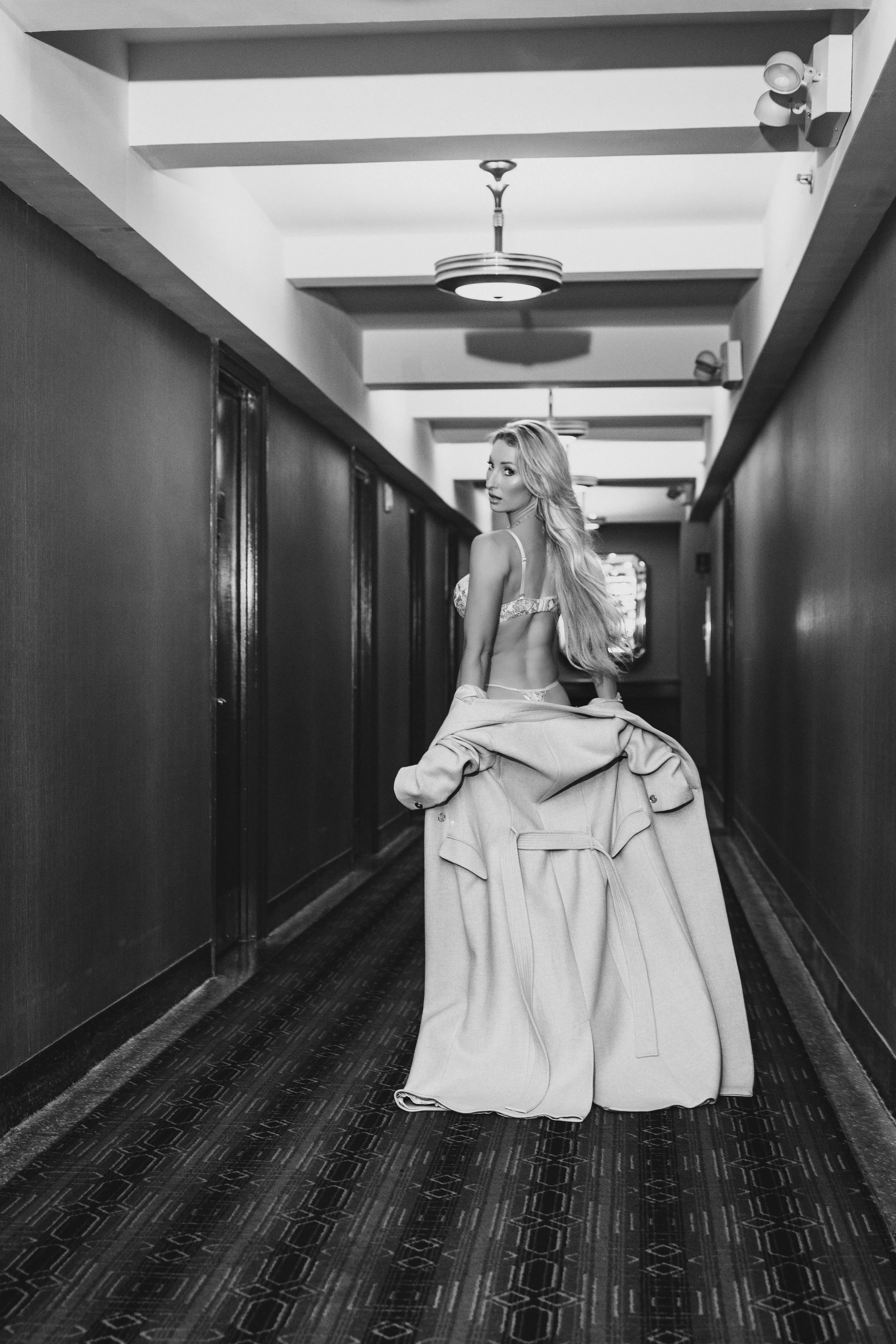 A woman with long, wavy hair standing in a hotel hallway, wearing lingerie and holding a coat, in black and white.