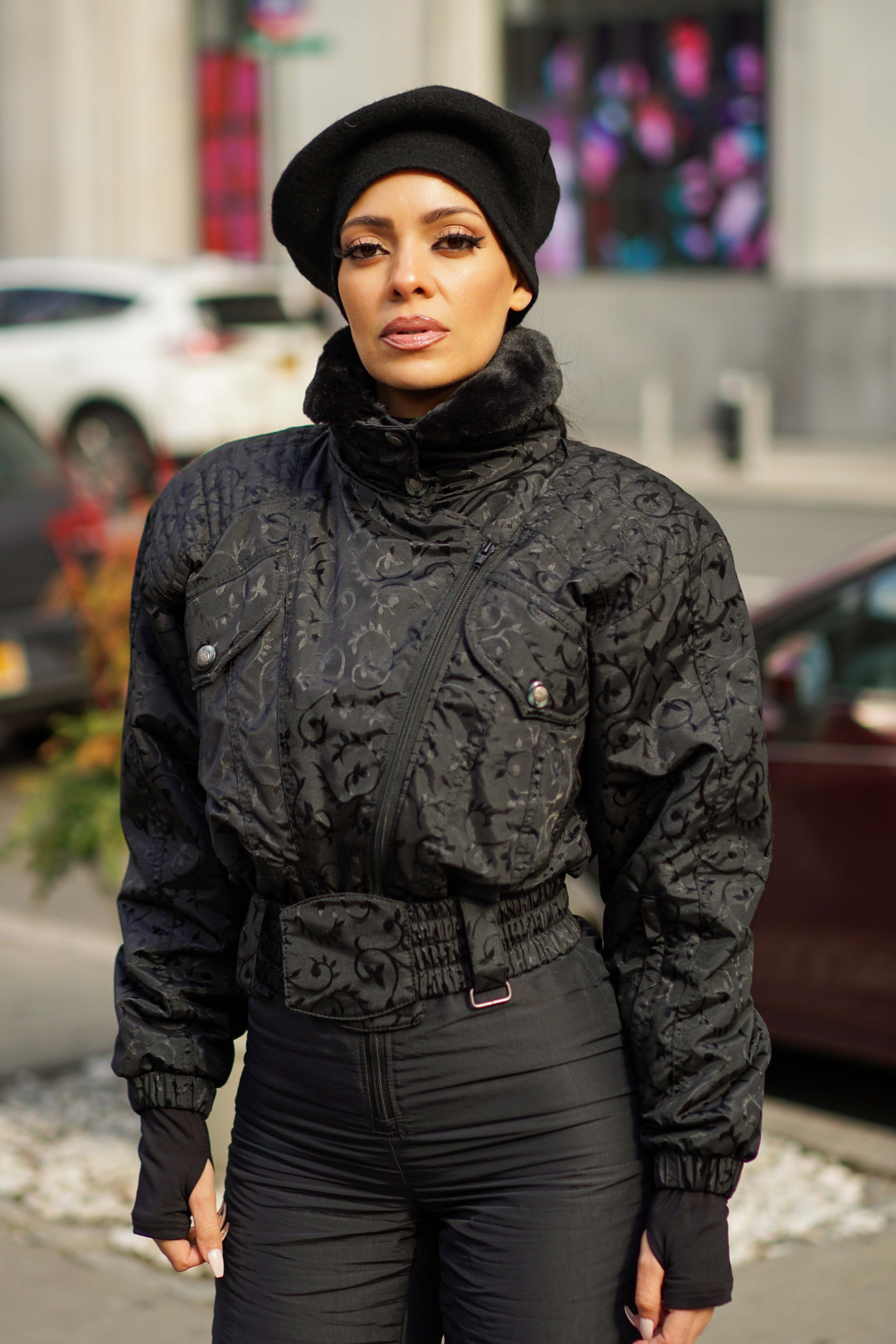 A woman wearing a black beanie, a black patterned jacket with a fur collar, and black pants, standing outdoors in an urban area with parked cars and a building with colorful artwork in the background.