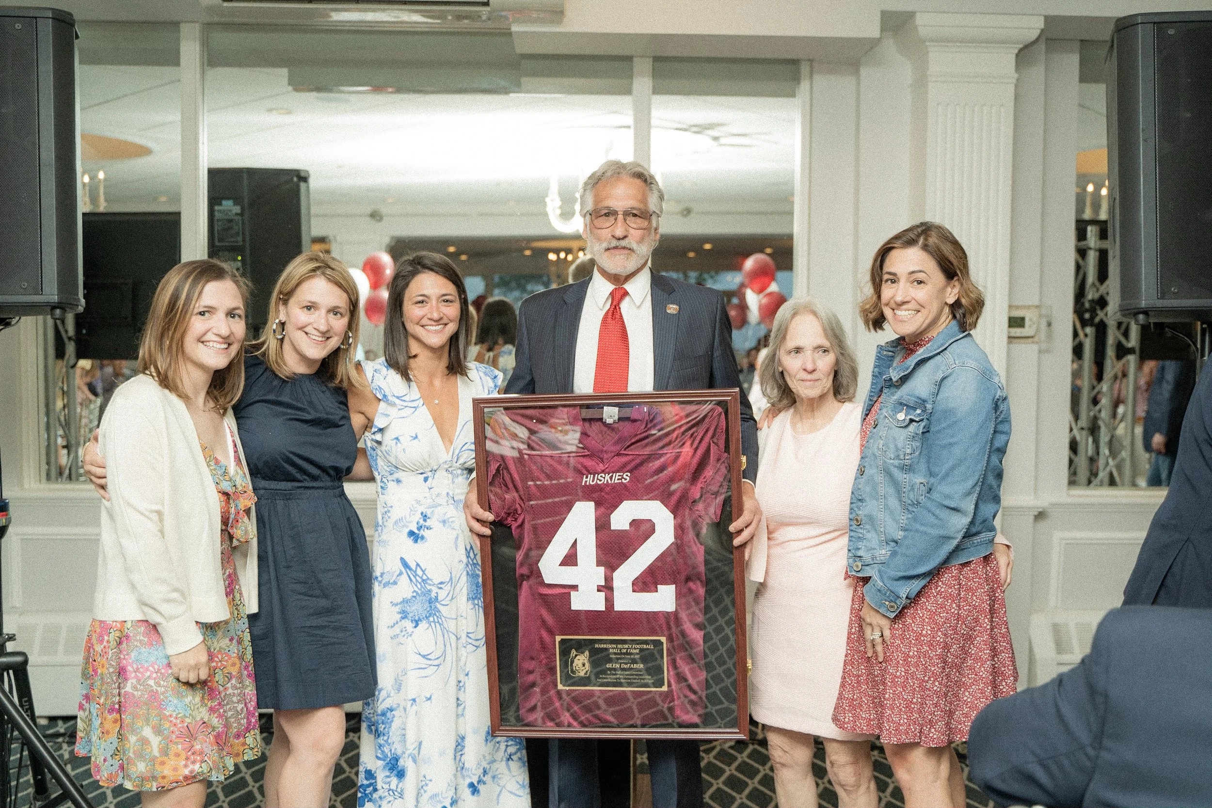 Group of six people standing in a room, smiling, with a framed jersey displaying the number 42 and a plaque in front of a man in a suit and tie. The background includes balloons and decorations.