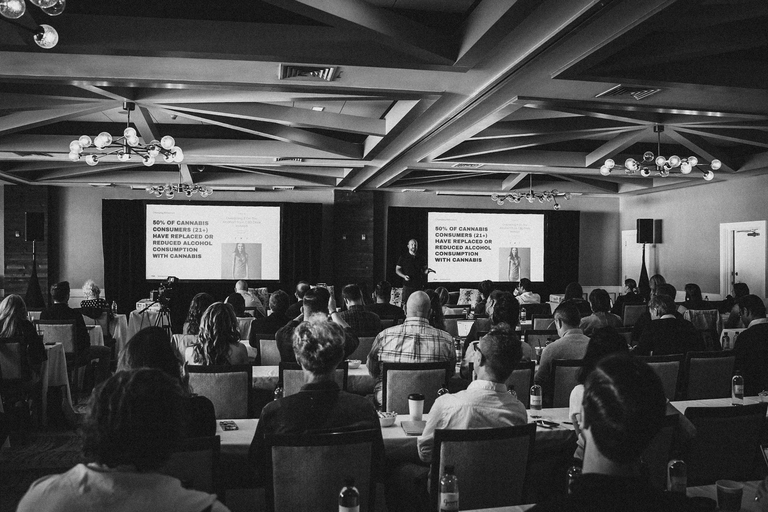 A black and white photo of a conference room filled with attendees listening to a speaker. The speaker is standing in front of two large screens displaying a presentation slide that reads, "50% of cannabis consumers (21+) have replaced or reduced alc
