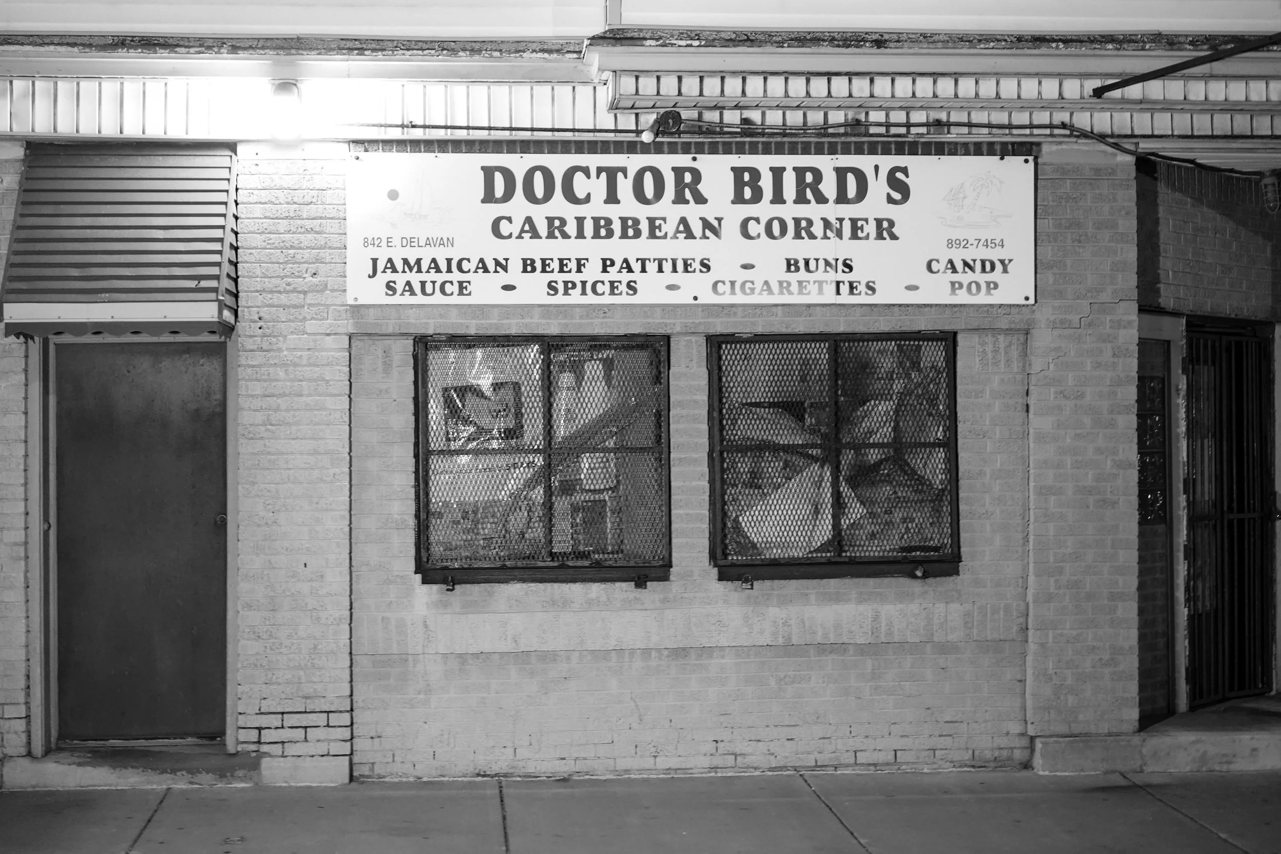 Black and white photo of a storefront with a sign reading 'Doctor Bird's Caribbean Corner' in Buffalo, New York offering Jamaican beef patties, buns, candy, sauce, spices, cigarettes, and pop. 